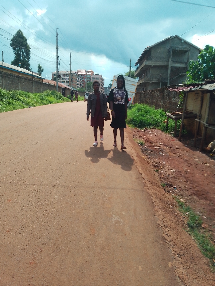 Two young women are walking down a paved road, away from the camera, in Rungiri, Kenya. They are both wearing casual clothing and one is carrying a handbag. There are several buildings visible in the background, including a large apartment building and a two-story house with a fence. The road is lined with vegetation on both sides, and the ground along the side of the road appears to be dirt. The sky is partly cloudy.
