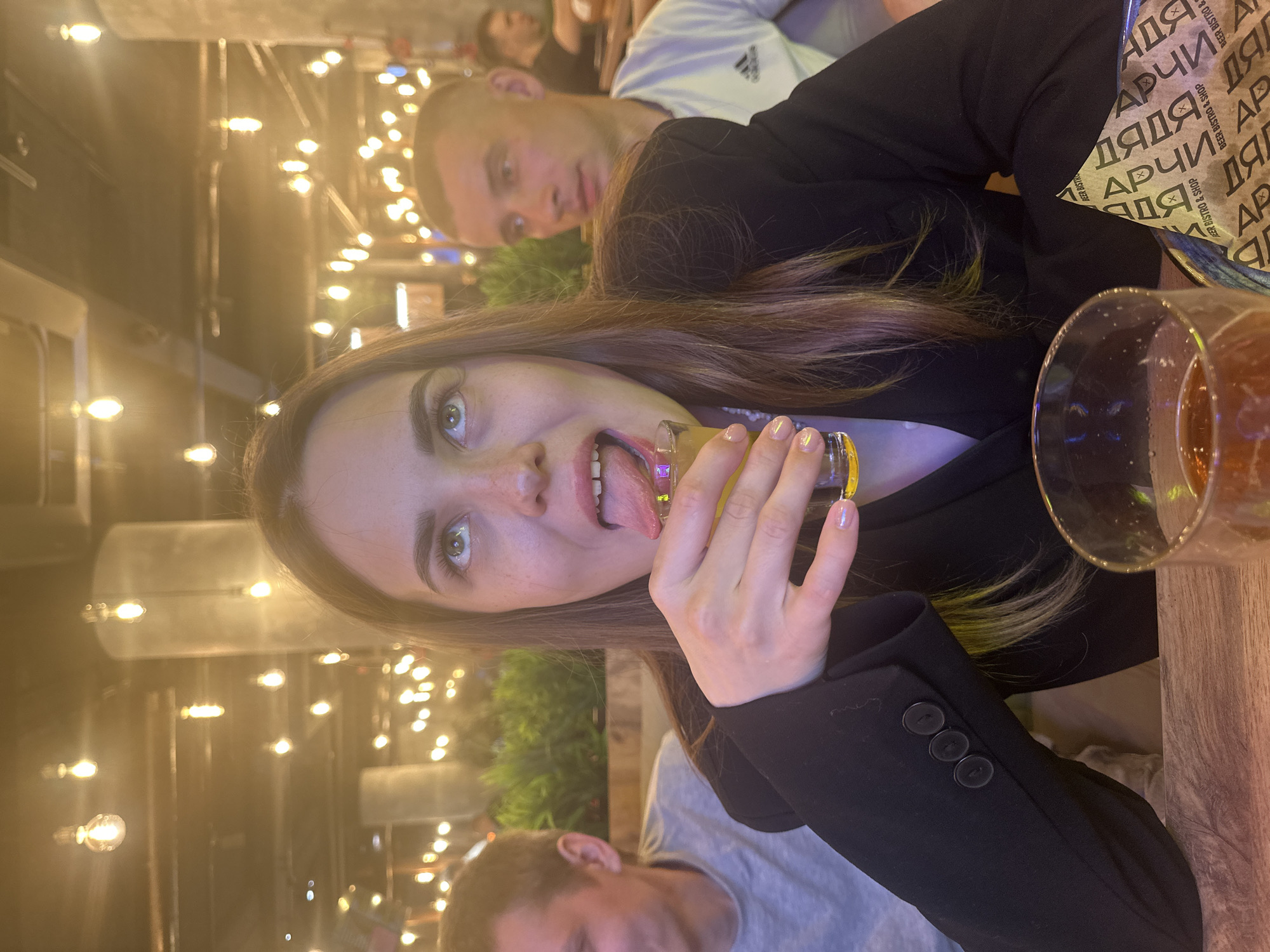 Here is a detailed description of the image:
**Scene:**
The image shows a young woman in what appears to be an indoor establishment, likely a bar or restaurant. The location is implied to be Perm, Russia due to the "ДЯДЯ АРЧИ" (Uncle Archi) branding on the packaging, a known establishment in that city. The lighting is warm and somewhat dim, with numerous decorative light bulbs visible in the background.
**People:**
* **Main Subject:** A young woman is the primary focus, taking a selfie with a shot glass to her lips, tongue extended.
* **Secondary Subjects:** Two men are partially visible in the background. One is directly behind the woman, looking into the camera. The other man is in the foreground and blurry.
**Objects:**
* Shot glass: The woman holds a filled shot glass to her lips.
* Drink: A larger glass of beer is on the right side of the image.
* Black Blazer: The woman is wearing a black blazer.
* Table: The drinks are resting on a wooden table.
* Branding: There is branded packaging with "ДЯДЯ АРЧИ" visible on the table.
* Lighting: Numerous decorative light bulbs provide warm lighting.
**Actions/Interactions:**
* The woman is taking a selfie with a shot glass to her lips, tongue extended, suggesting a playful or celebratory moment.
* The man in the background is looking at the camera.
**Environment:**
The setting appears to be a trendy bar or restaurant, given the lighting and decor.