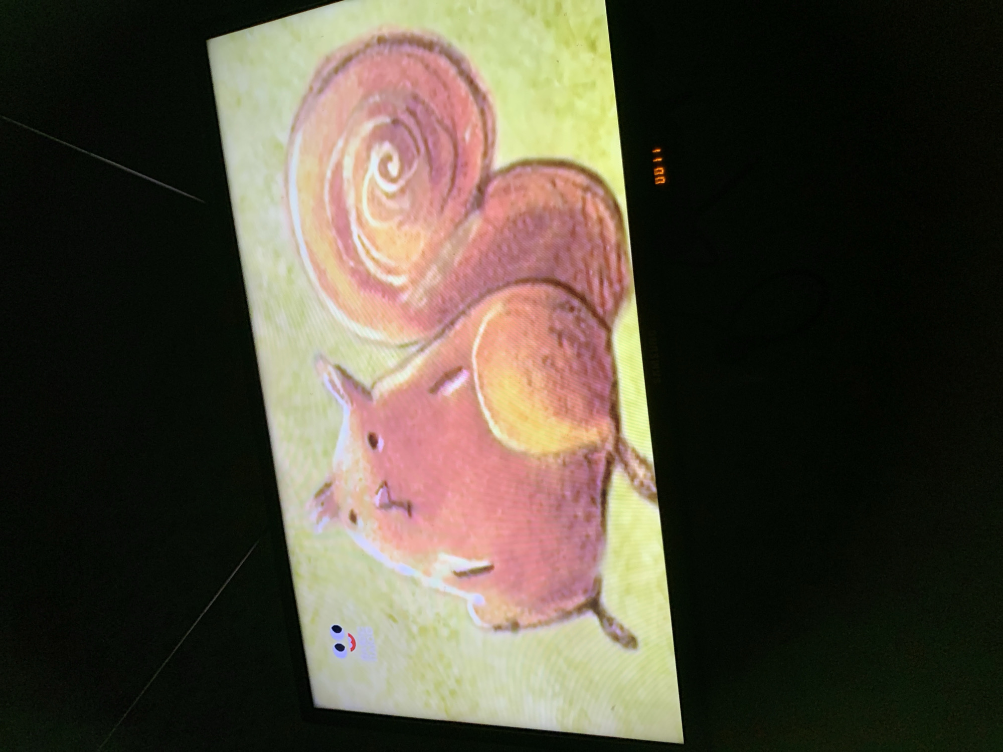 A television screen displays a cartoon illustration of a pink squirrel with large ears and a bushy, spiraled tail. The squirrel is depicted in a standing or sitting pose against a light green background with a textured pattern. In the upper left corner of the screen, there is a small, stylized blue face with two dots for eyes and a red curved line for a mouth. Along the bottom edge of the screen, the word "SAMSUNG" is visible, and to its right, a digital display shows "00 11" in orange LED lights. The image is captured in a dark room, suggesting the television is the primary light source. The location in Lymanka, Ukraine is not discernible from the visual information provided.