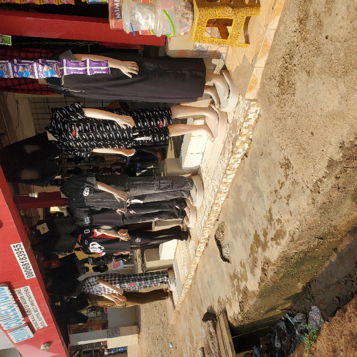 The image depicts a street scene in Eziobodo, Nigeria, specifically in front of a retail shop named "EMMA COLLECTIONS." The shop has a red facade with white lettering and displays contact information, including a phone number "08068183955" and a description: "DEALERS IN ALL KINDS OF WEARS LIKE LADIES, GENTS, BELT, WRISTWATCHES."
Six headless mannequins are positioned along a tiled sidewalk directly in front of the shop, displaying various clothing items. From left to right: one mannequin wears brown trousers (partially visible); another wears black and white checkered pajamas; a third wears a black t-shirt with a white "20" logo and black trousers; a fourth displays a dark short-sleeved shirt over a black t-shirt with dark cargo pants; a fifth wears a black short-sleeved button-up shirt with white logos and matching shorts; and the sixth wears a long black dress. Items, possibly more clothing, hang from the shop's awning. A string of purple-packaged snacks is suspended near the rightmost mannequin.
Adjacent to the sidewalk is a concrete drainage ditch filled with refuse, including plastic bottles, bags, and muddy water. A wooden plank bridges a section of this ditch on the left side. Beyond the ditch is an unpaved, dusty ground area. On the far right, a yellow plastic stool holds stacked clear plastic containers, the bottom one containing what appears to be snacks. The scene is illuminated by bright daylight. No human individuals are visible.