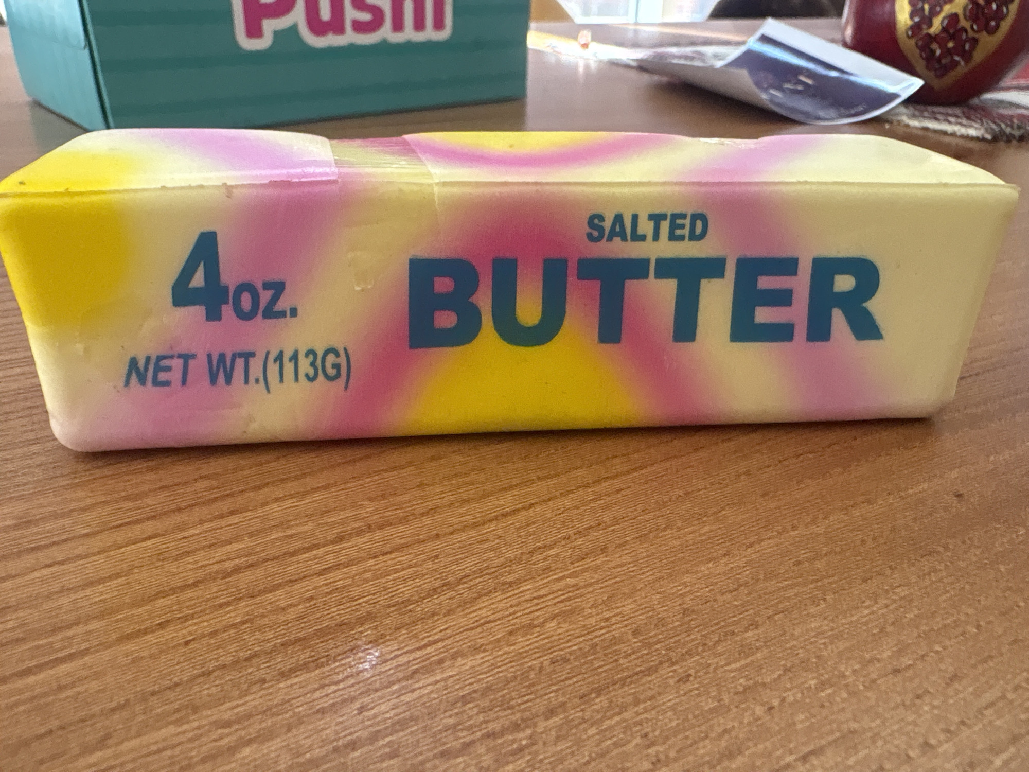 A close-up, eye-level shot shows a stick of butter with a tie-dye wrapper of yellow, pink, and white. The butter is on a wooden table. The word "SALTED" is printed above the word "BUTTER" in large, teal capital letters. Below "BUTTER," "4oz." and "NET WT.(113G)" are printed in the same color. In the background, a teal box with "Pushi" printed on it in pink letters is visible. The image was taken in Georgiyevsk, Russia.