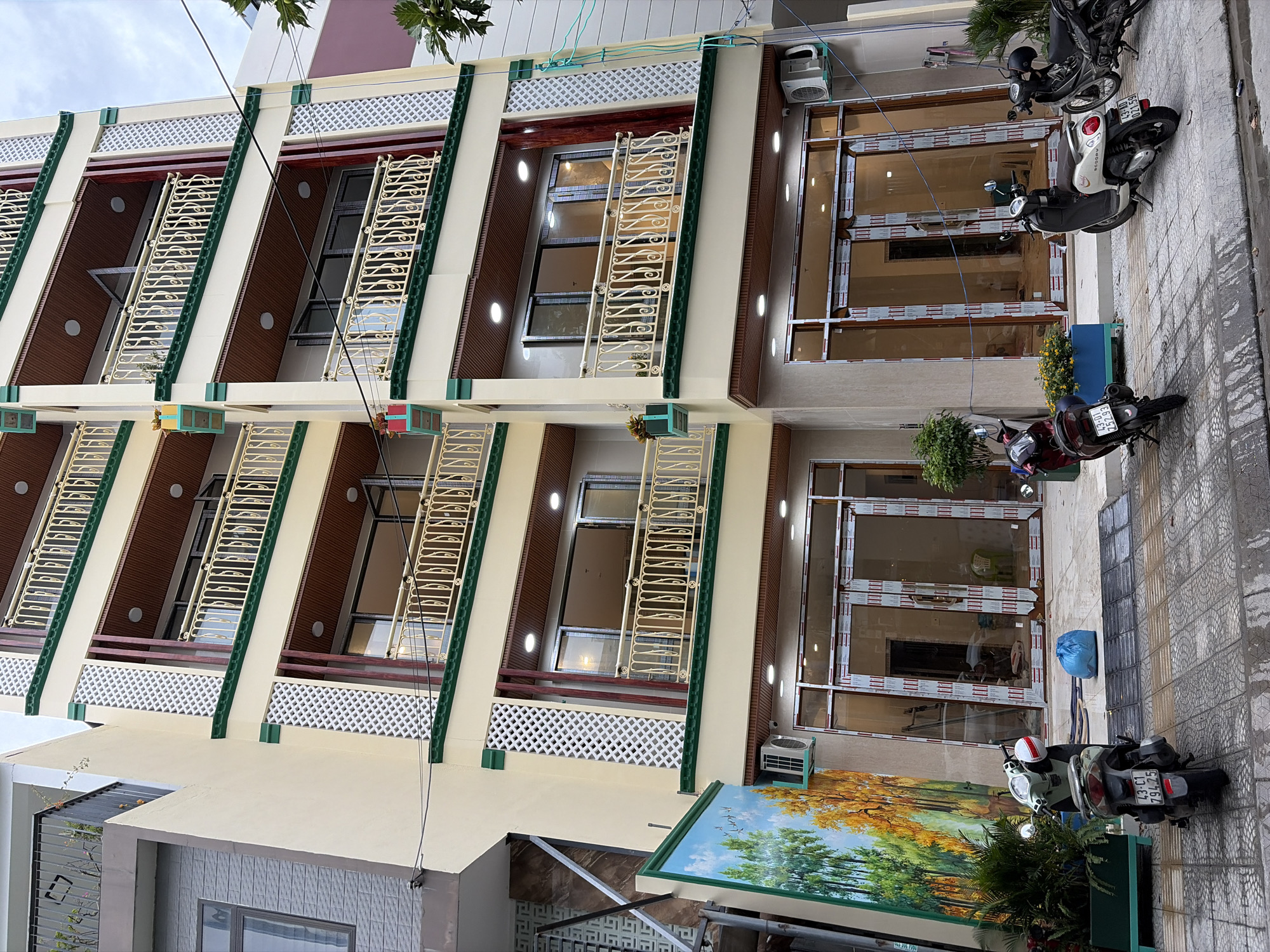 This is a full, eye-level, medium shot of a modern, multi-story building with a light yellow facade, located in the city of Đà Nẵng, Vietnam. The building has multiple balconies with decorative metal railings and green trim. The ground floor features large glass storefronts. Several motorcycles are parked on the sidewalk in front of the building. A large mural depicting trees and a flock of birds adorns the left side of the building's exterior. The sky is overcast. There are no people visible in the image.