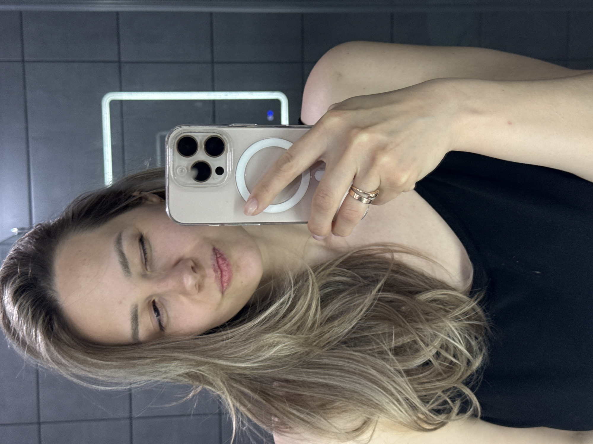 The image is a selfie taken in a bathroom, possibly in Chaikovsky, Russia. A woman with shoulder-length blonde hair is holding an iPhone with a clear case, which features a MagSafe ring. She is wearing a black top and has two rings on her finger. The background shows a grey tiled wall with a reflective surface, possibly a mirror and a rectangular light fixture. The woman is winking.