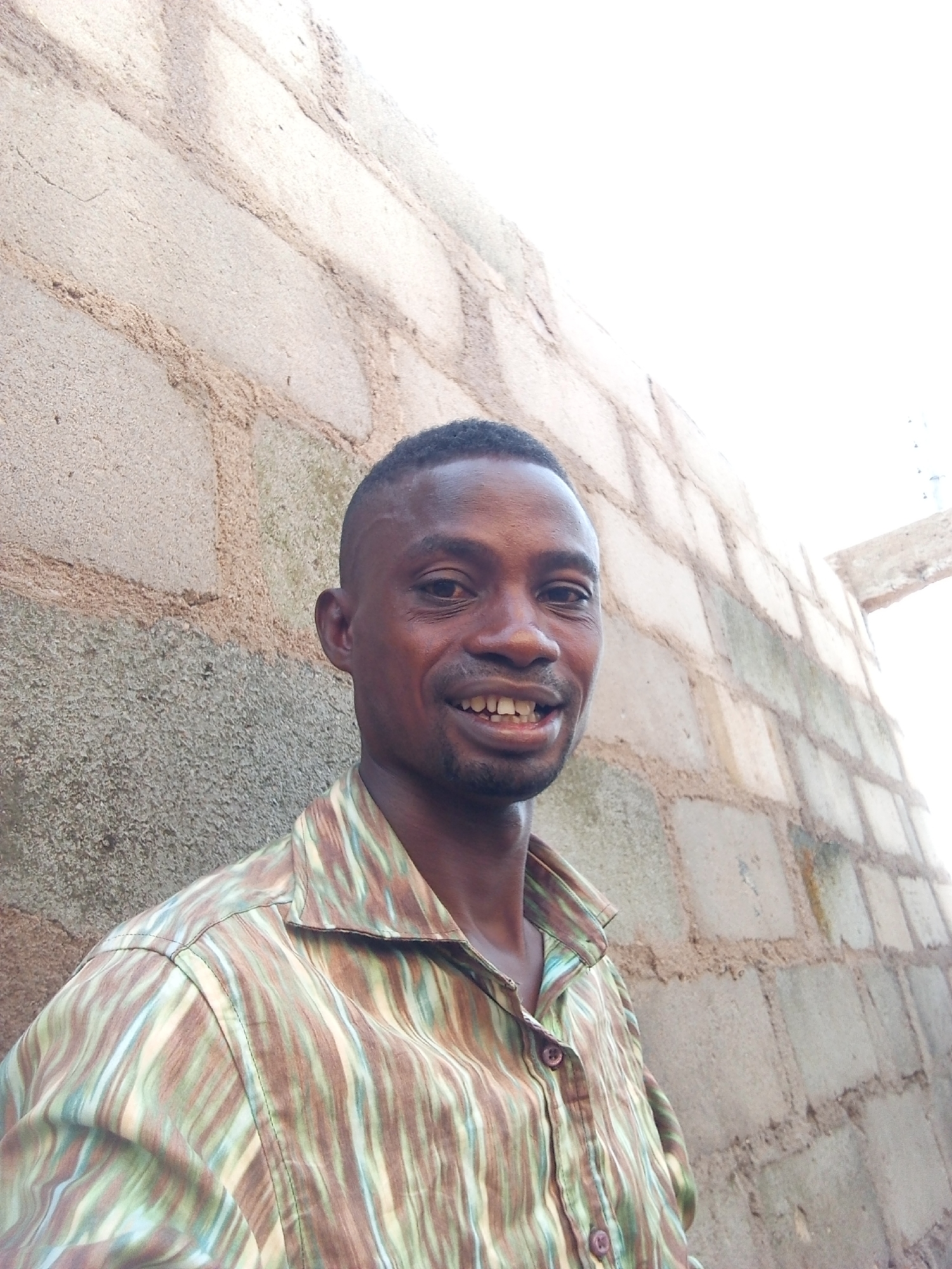 The image captures a portrait of a man, likely of African descent, smiling and looking directly at the camera. He has dark skin, short dark hair, and a neatly trimmed beard. His expression is friendly and cheerful, with his mouth open in a smile that reveals his teeth. He is wearing a collared, short-sleeved button-down shirt featuring a vertical striped pattern in shades of brown, green, and light beige or cream.
The background consists of a wall made of light-colored concrete blocks. The wall appears unpainted and has a rough, textured surface. Distinct mortar lines are visible between the blocks. There are also some darker, earthy-toned streaks or patterns on the wall, particularly noticeable in the upper left and central areas. The upper right portion of the image is brightly overexposed, suggesting strong sunlight and a clear, bright sky. This indicates the photo was taken outdoors during the daytime. The overall setting appears to be a simple, outdoor environment, consistent with the context of Adonkolo, Nigeria. There is no visible text in the image.