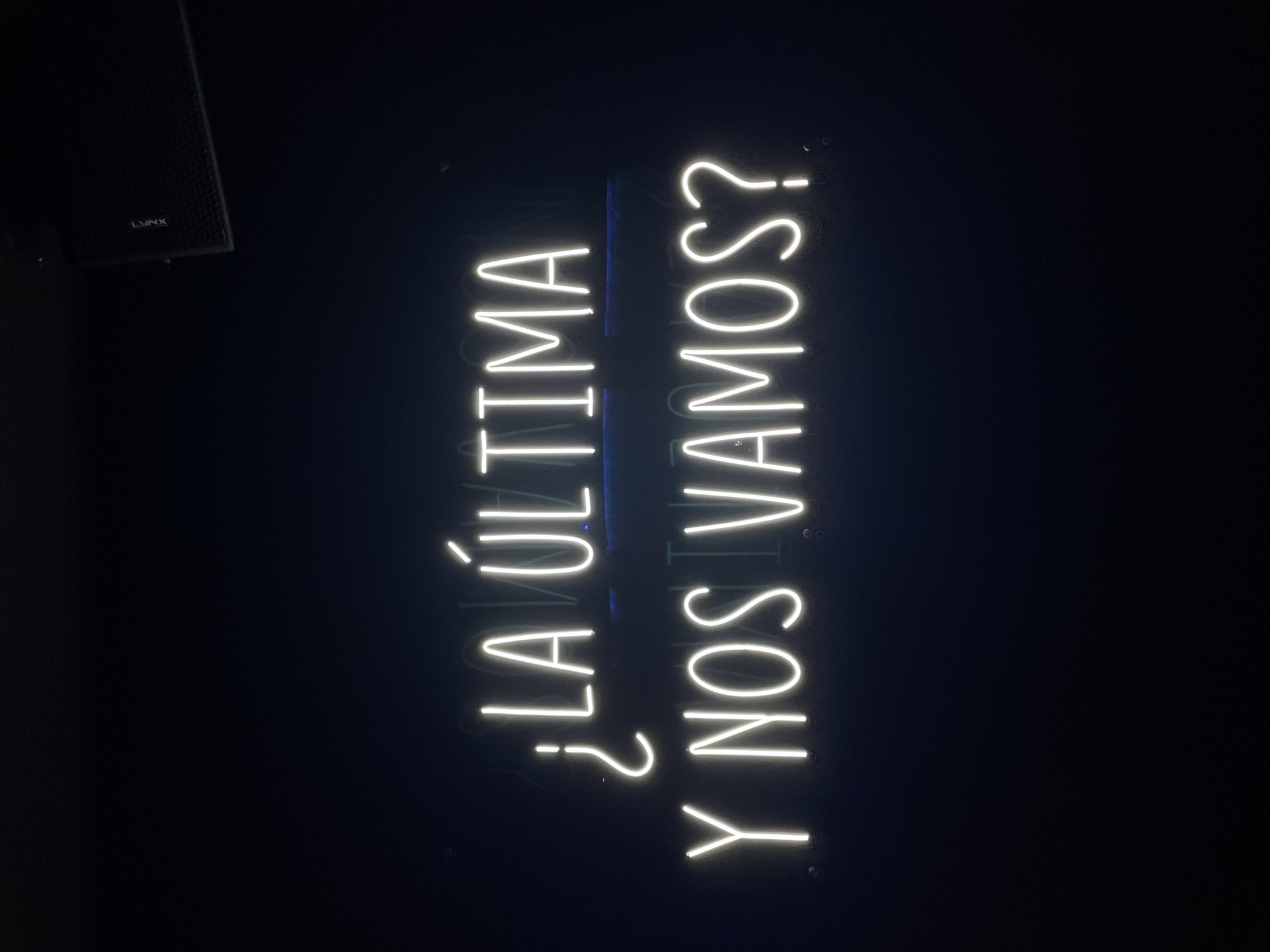 The image depicts a neon sign reading "¿LA ÚLTIMA Y NOS VAMOS?" (Is it the last one, and we leave?) vertically oriented against a dark background. A speaker labeled "LYNX" is visible in the upper left. The setting is implied to be Barcelona, Spain, due to the Spanish phrase. The environment appears to be a bar or club given the context of the phrase and the presence of the speaker.
