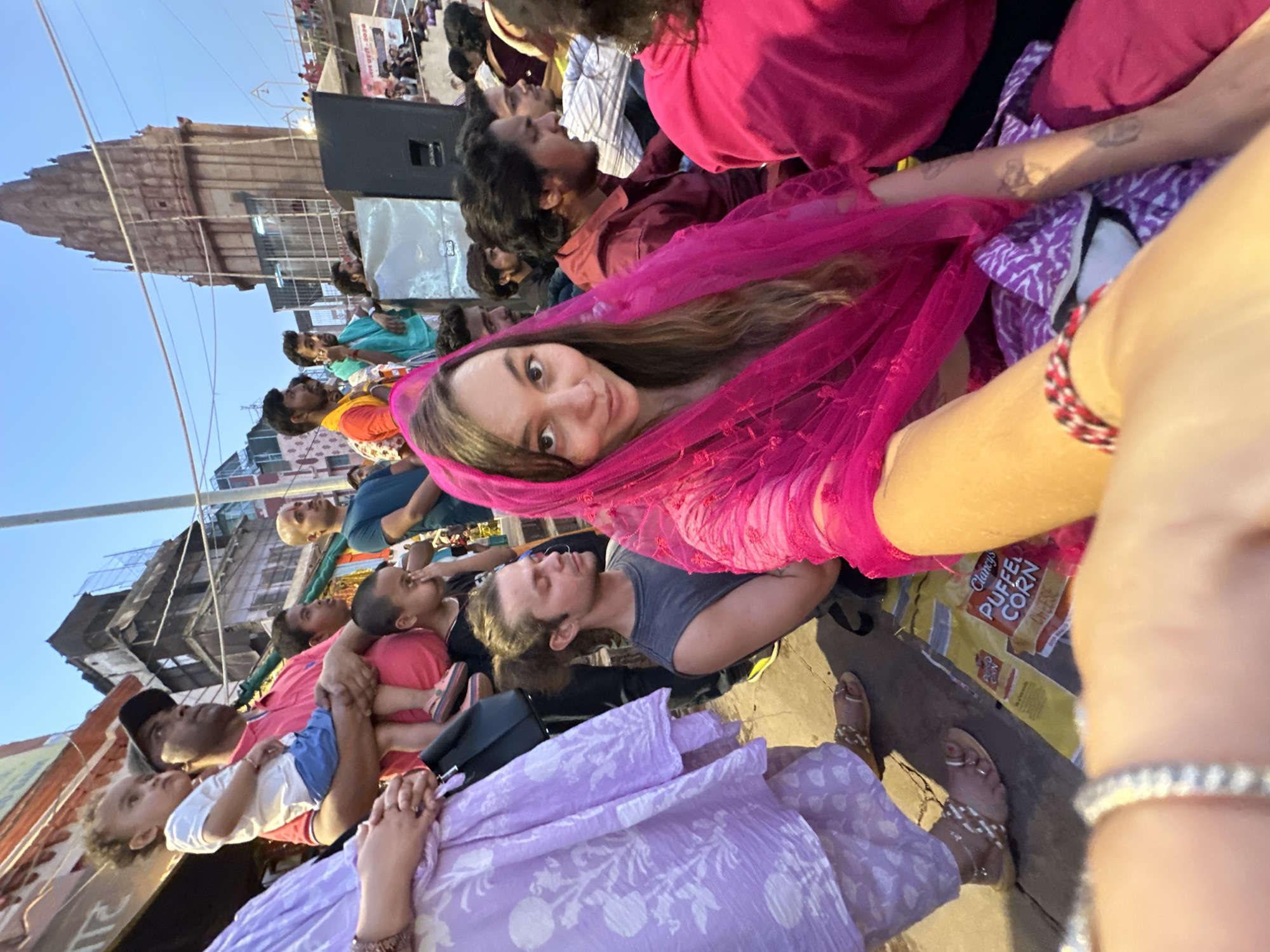 A ground-level, wide-angle, eye-level shot depicts a diverse crowd of people sitting on the ground in an outdoor urban setting, presumably in Varanasi, India, given the visible temple architecture in the background.
In the foreground, a woman is looking into the camera, draped in a bright pink, semi-sheer scarf or dupatta. Other people are visible around her, seated closely together. To the left, a person is wearing a light purple patterned outfit. Further back, one man holds a child, and various others are observing something in the distance, presumably a performance or event.
The setting appears to be near a temple or religious site, evidenced by the structure in the background. The sky is visible in the upper left, suggesting it's daytime. The light is somewhat harsh. Some people are sitting on what appears to be packaging or discarded items. There appears to be a speaker near the background temple.