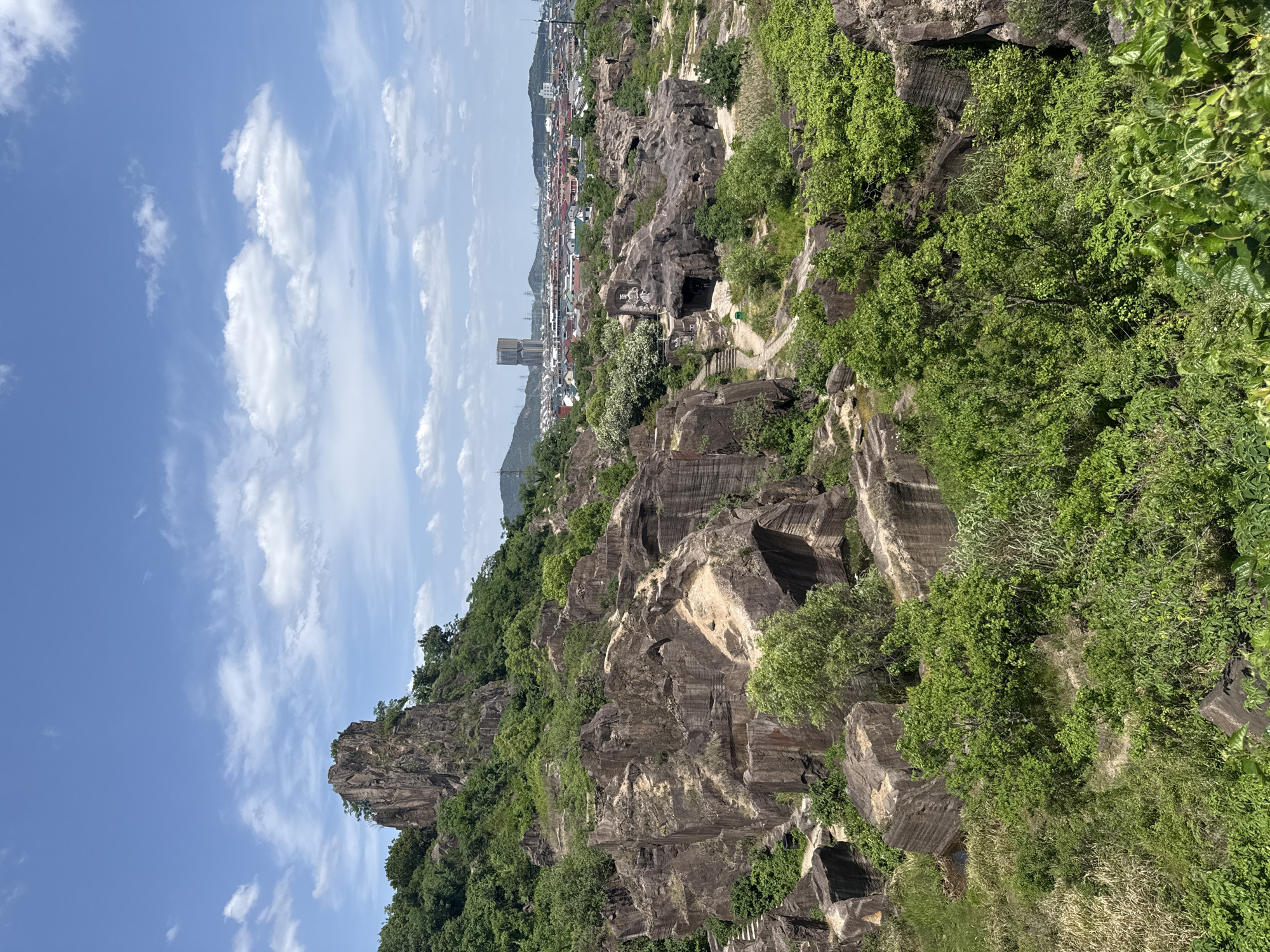 The image shows a view of the Anchang Stone Quarry in Keqiao District, China. It features the rocky, tiered landscape of the former quarry, with areas of green vegetation covering the stone. In the distance, a city skyline is visible with buildings and a single tower. The sky is blue with scattered white clouds. The image depicts a landscape that blends natural features with traces of urban development.