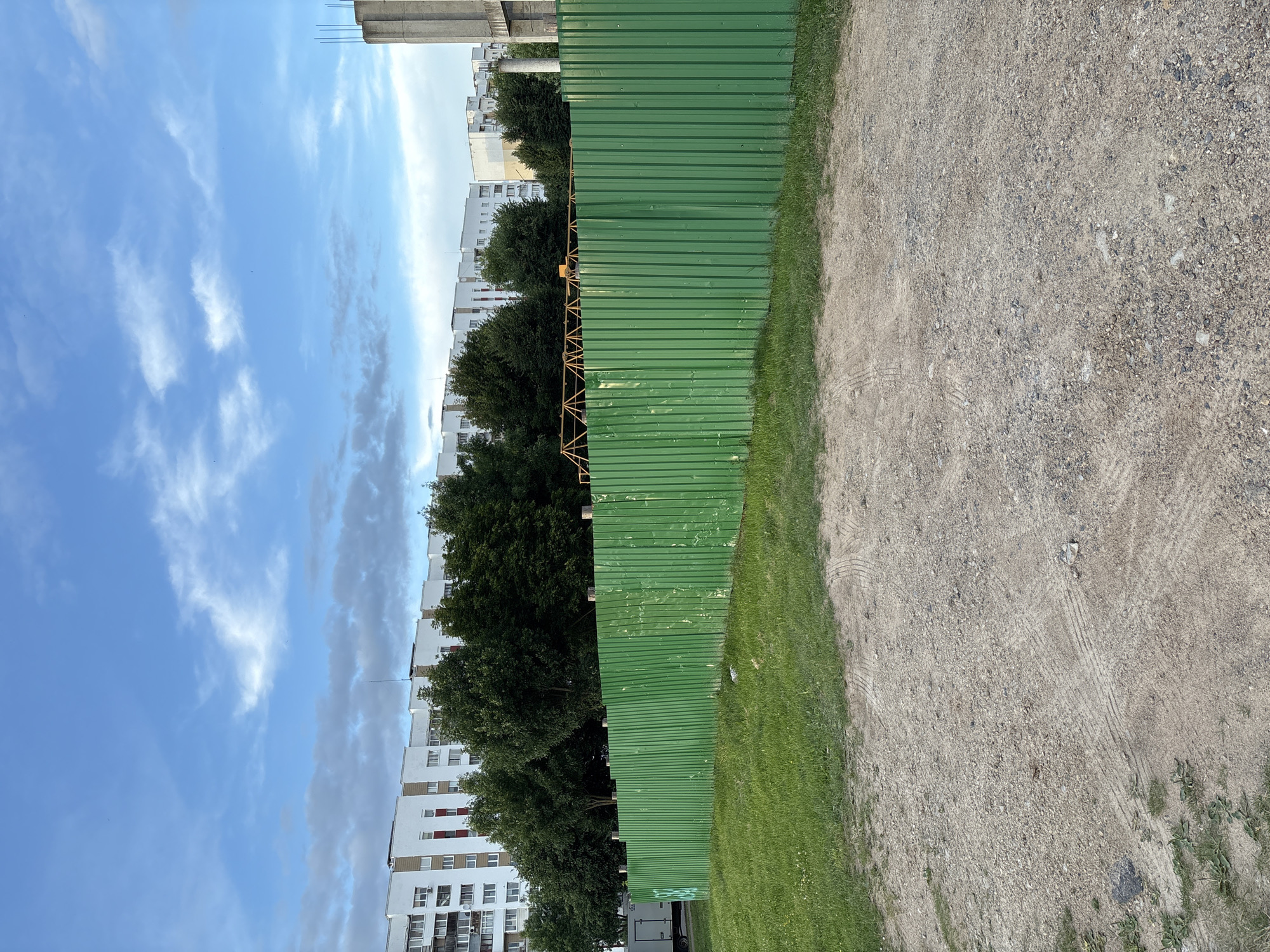 An outdoor, eye-level, medium shot captures a construction site in Vitsebsk, Belarus. A bright green corrugated metal fence stretches horizontally across the frame, dominating the foreground. Behind the fence, a line of lush green trees forms a dense barrier. Beyond the trees, a row of multi-story apartment buildings, characterized by their white facades and numerous windows, extends into the distance. The sky above is a mix of blue with scattered white clouds. In the far right of the image, the top of a concrete structure, possibly part of a new building, is visible. The ground in front of the fence is a mix of gravel and dirt, with tire tracks visible in the foreground. A small white trailer is parked to the left of the frame, partially obscured by the fence. The scene appears to be at dusk.