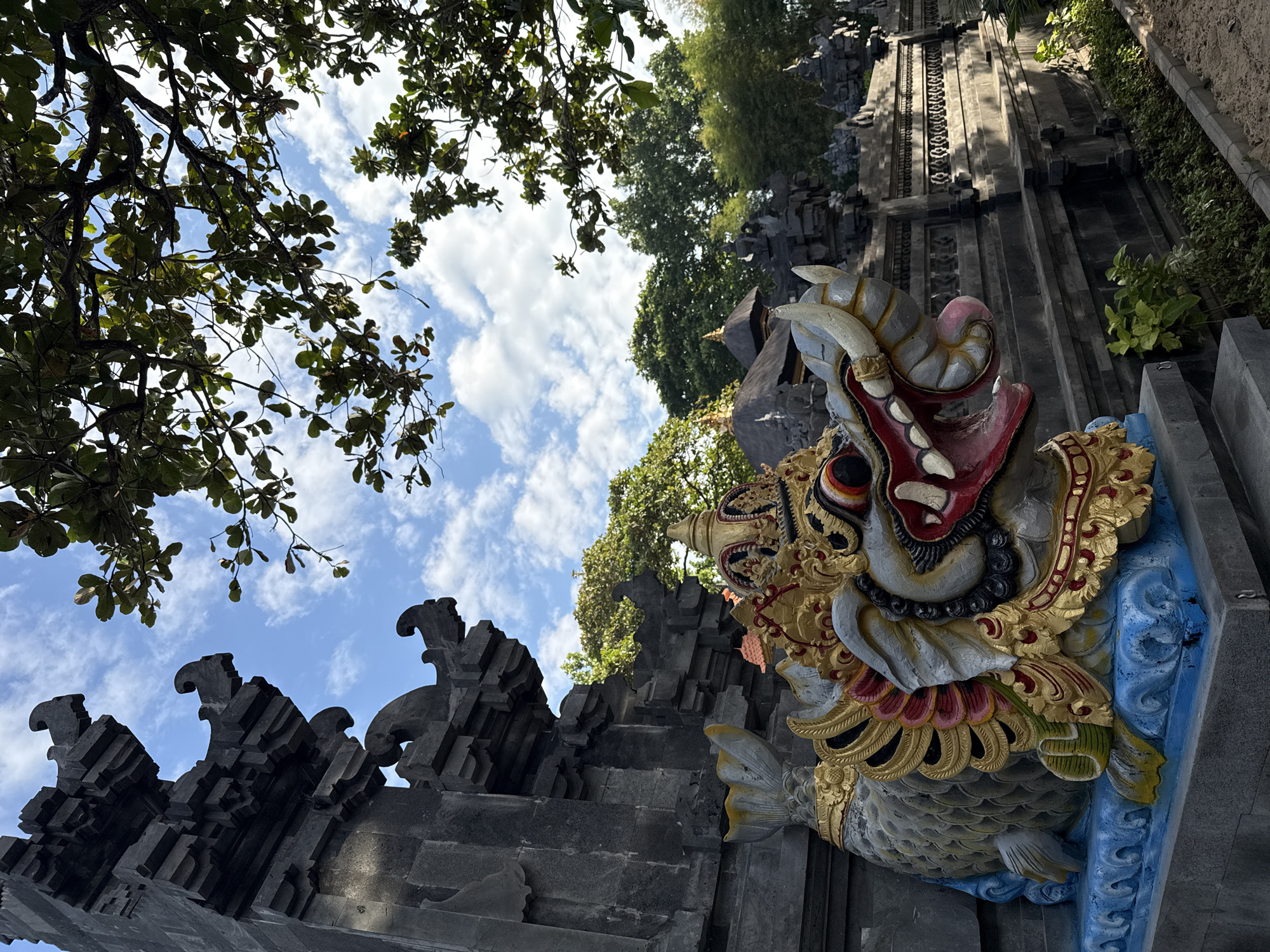 This image depicts a detailed, multi-colored statue of a mythical creature, likely a Makara, positioned prominently in front of traditional Balinese temple architecture under a clear sky. The statue, occupying the lower-left to central foreground, features a fish-like body with gray scales and blue fins, resting on a blue sculpted base. Its head is intricately adorned with gold, red, black, and white paint, showcasing a crown, prominent tusks/horns, fangs, a red tongue in an open mouth, and decorative elements around its neck.
Behind the statue and extending along the left side of the frame is a towering structure made of dark gray, possibly volcanic, stone, carved with traditional Balinese motifs. Further into the background, additional dark stone temple walls and structures with dark, possibly thatched, tiered roofs are visible, surrounded by lush green trees and foliage. The upper portion of the image is dominated by a bright blue sky with scattered white cumulus clouds, visible through the canopy of dark green leaves and branches that partially frame the top and right sides of the scene. The ground in the lower right shows a sandy or dirt path adjacent to a small bed of green plants. No people are visible. The scene is illuminated by natural daylight, casting subtle shadows. This location is within Kuta Selatan, Indonesia.