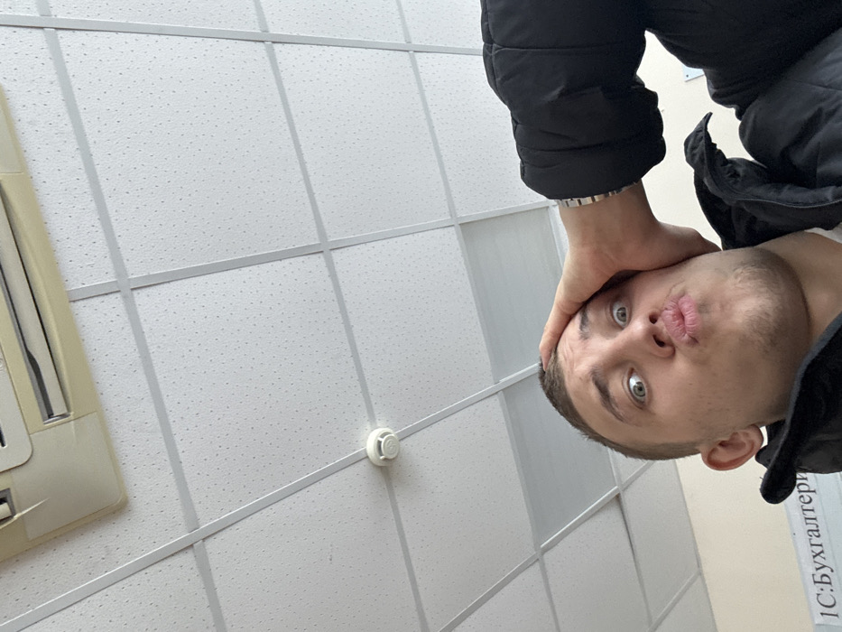 A young male is depicted in an indoor setting, captured from a low-angle selfie perspective. He has short, dark hair, wide-open eyes, and pouted lips, looking directly at the camera. His left hand is placed on his forehead, fingers splayed. He is wearing a dark puffer jacket, with a metallic-banded watch visible on his left wrist. The overhead environment consists of a white dropped ceiling with acoustic tiles arranged in a grid pattern. An off-white air conditioning unit is partially visible in the upper left corner of the ceiling, and a white, round smoke detector is centrally mounted on a ceiling tile. In the lower left background, partially visible Cyrillic text on a light-colored wall reads "1С: Бухгалтерия," which translates to "1C: Accounting." This detail indicates the location is an office, classroom, or training facility related to business or accounting in Chaikovsky, Russia.