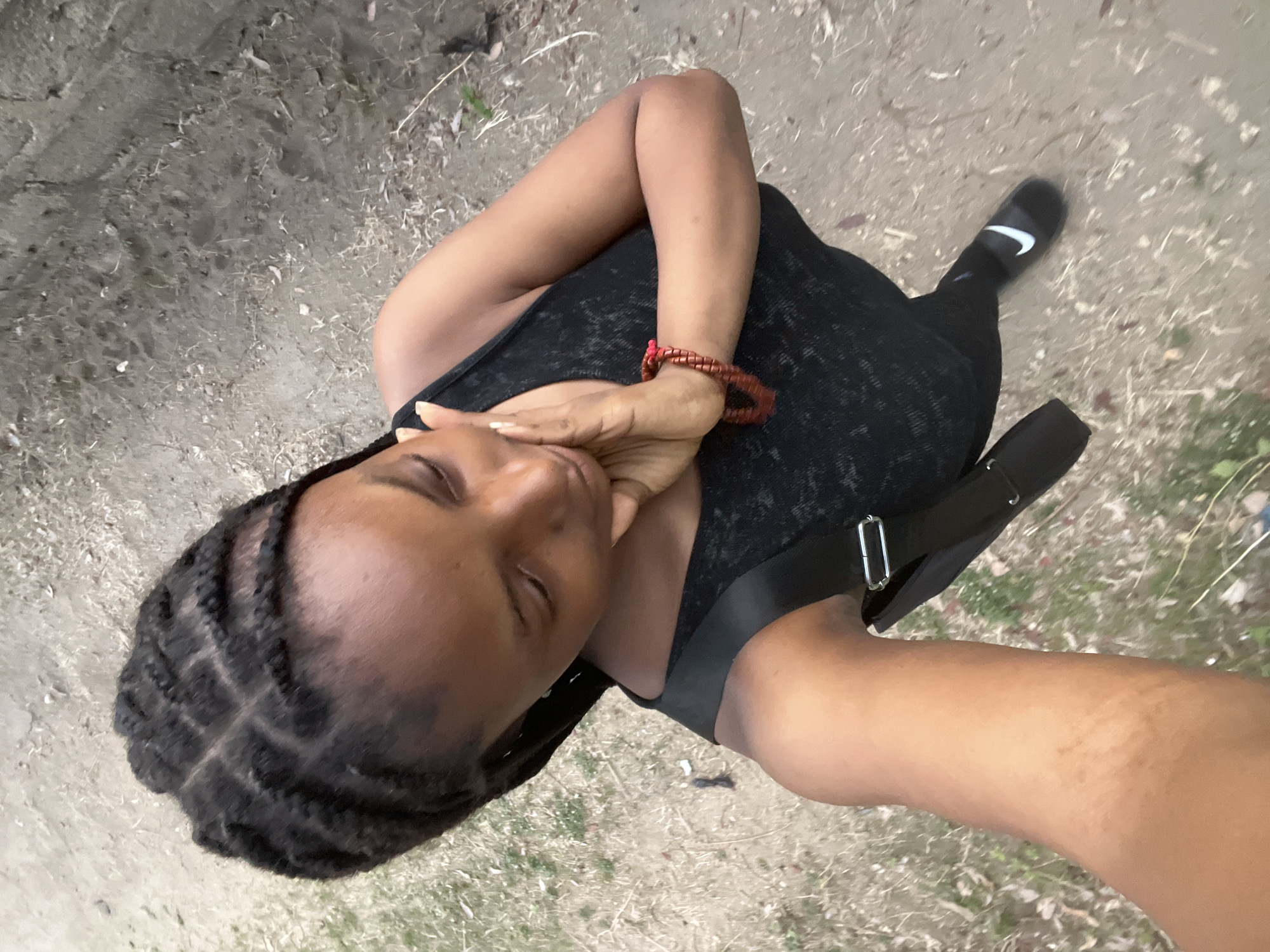 The image is an upward-angled selfie of a Black woman standing on a dirt path. She is wearing a black tank top, black leggings, and black Nike slides. She also wears a red beaded bracelet on her left wrist. A black bag with a silver buckle is strapped across her body. She is touching her face with her hand. Her hair is styled in braids. The background is a dirt path with some vegetation. Given the presence of dry, earthy ground and sparse vegetation, the location could be in a less urbanized part of Lokoja, such as near the edges of the city or in a park-like environment.
