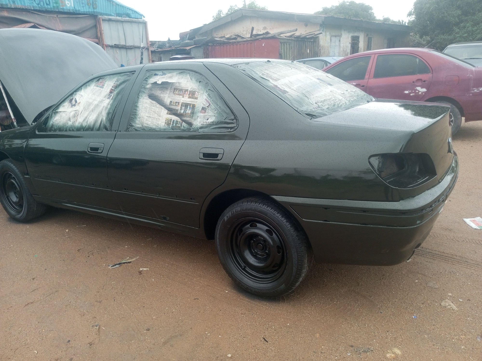 In New Stadium, Nigeria, a dark green sedan is parked on a dirt ground. The car's hood is open, and its windows are covered with newspapers, suggesting it is being prepared for painting or has recently been painted. The car appears to be in a state of repair or maintenance. In the background, a red sedan is also visible, along with some dilapidated structures and a shipping container. The ground is covered with sand and debris, indicating an outdoor, possibly informal, workshop or storage area. There are no people visible in the image.