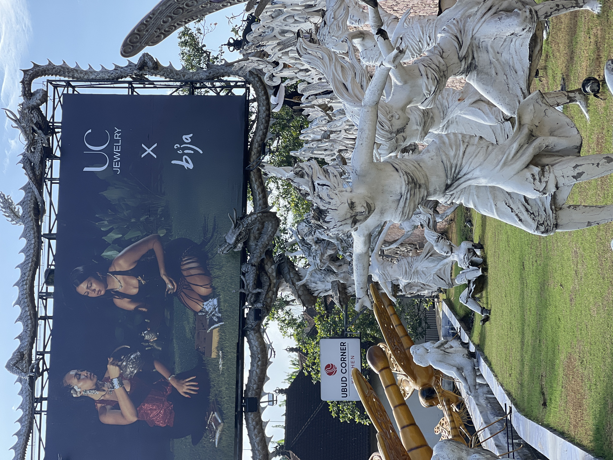 The image displays a billboard advertisement for "UC JEWELRY X bija" situated above a large outdoor sculpture garden in Sukawati, Indonesia. The billboard features two women in traditional attire, with the text "UC JEWELRY" and a stylized "X" and "bija" logo. In the foreground, the sculpture garden is filled with numerous white, distressed statues depicting human figures in dynamic poses, some appearing to be dancing or struggling. Behind the statues, a sign reads "UBUD CORNER KITCHEN", indicating the location. The environment is outdoors with green grass and trees visible in the background. A large, dragon-like sculpture is mounted above the billboard, adding a fantastical element to the scene.