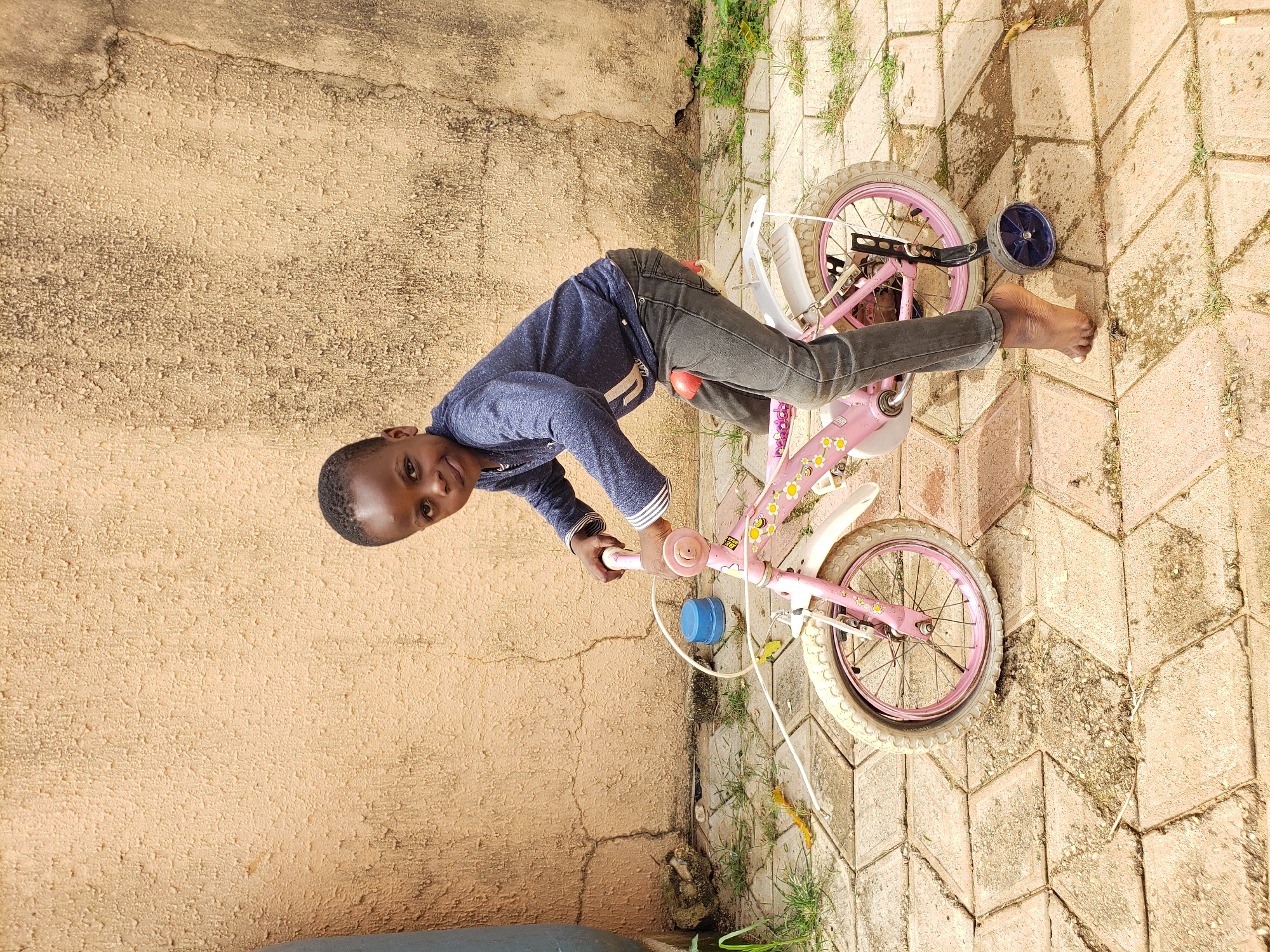 A young Black child with short hair and dark eyes is sitting on a pink bicycle with training wheels. The child is wearing a dark blue long-sleeved shirt and dark gray pants, and is barefoot. The bicycle has a white fender over the back wheel and a blue cap on the handlebars. The child is looking at the camera and smiling.
The scene takes place outdoors in Sabon Tasha, Nigeria. The background consists of a textured, pale orange wall. In front of the wall is a paved ground surface made of interlocking diamond-shaped bricks. Some patches of green grass and a few fallen yellow leaves are visible on the ground. A blue cylindrical object is resting on the ground to the left of the bicycle. The lighting suggests it is daytime.