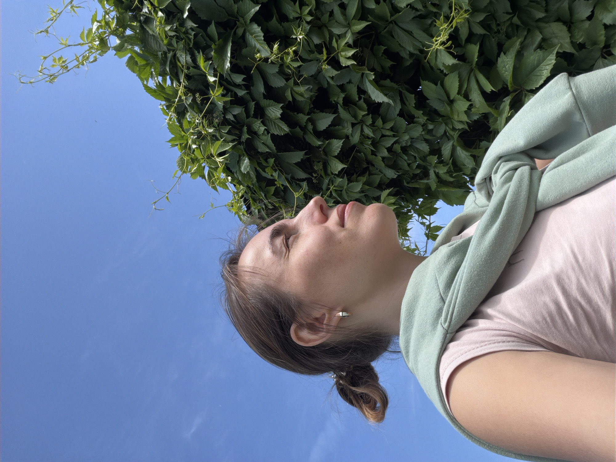 A young adult woman is depicted in profile, facing right with her eyes slightly closed and a subtle smile, illuminated by bright daylight. She has light brown hair secured in a low bun and wears a small, angular metallic earring on her visible ear. Her attire consists of a light pink short-sleeved top, with a light green sweatshirt casually draped over her shoulders. The background features a clear blue sky with minimal cloud cover in the upper and left sections. To her right, a dense, vertical expanse of dark green foliage, characteristic of a climbing vine, dominates the space, with lighter green new growth and tendrils extending into the sky. The environment is outdoor and sunny. No visual identifiers within the image confirm the specific location as Sarapul, Russia.
