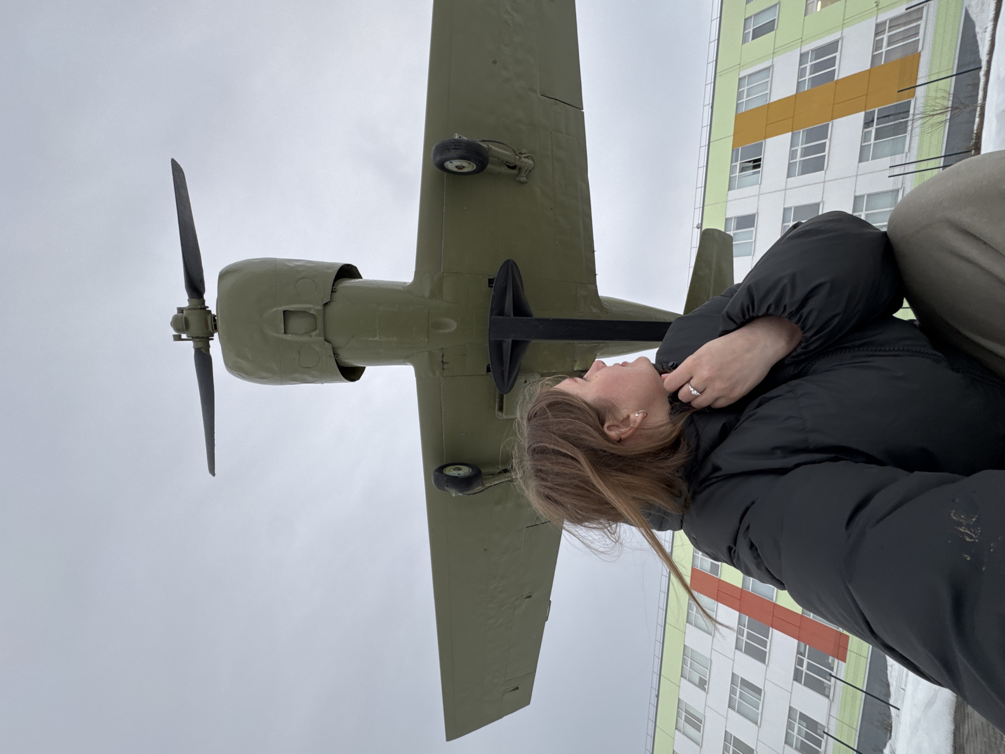 An outdoor photograph captured in Ферма, Russia, depicts a female individual in the foreground, looking upwards at a static aircraft display. The individual, positioned in the lower-left, is seen in profile from the chest up, with light brown hair and wearing a dark puffer jacket. A silver-colored ring is visible on her right ring finger. Above her, a large, olive-green, single-engine propeller aircraft is mounted on a black stand, viewed from its underside, showcasing its wings, propeller, and landing gear wheels. In the background, a multi-story building with white walls, multiple windows, and vertical accent panels in lime green and orange/gold is visible. Patches of snow are present on the ground. The sky is uniformly overcast.