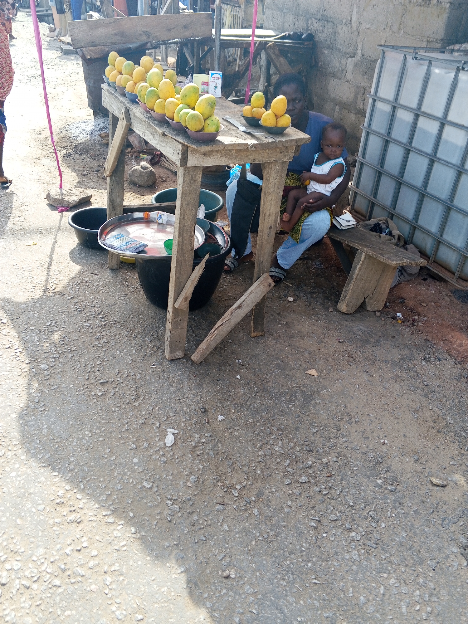 An eye-level, vertical shot captures a bustling outdoor market scene in Adankolo, Nigeria, centered on a woman vendor and her baby. In the foreground, a rustic wooden table displays numerous bright yellow and green mangoes, neatly arranged in small bowls. A knife rests on the table alongside the fruit.
The vendor, a woman in a blue top and light-colored trousers, is seated on a low wooden bench behind the table, holding a baby dressed in a white sleeveless top. Her gaze is directed towards the front, suggesting she is attending to potential customers. To her right, a weathered concrete block wall rises, partially obscured by a large, white translucent water storage container secured within a metal cage.
On the ground around the table, several black plastic tubs are visible. One tub holds a metal tray with a partially consumed Fanta bottle, several clear sachets (possibly water or juice), and a green cup. A pink pole leans against a rock on the left side of the frame, possibly part of another stall or support structure. The ground is a rough, gravelly dirt path, typical of informal market environments.
The bright, direct sunlight indicates it is daytime, casting clear shadows and highlighting the textures of the ground and objects. Visible text includes "BABYTALK BABYTEA" on a container placed near the mangoes and "FANTA" on the soft drink bottle. The image portrays a slice of everyday life, blending commerce with familial care in a vibrant outdoor setting.