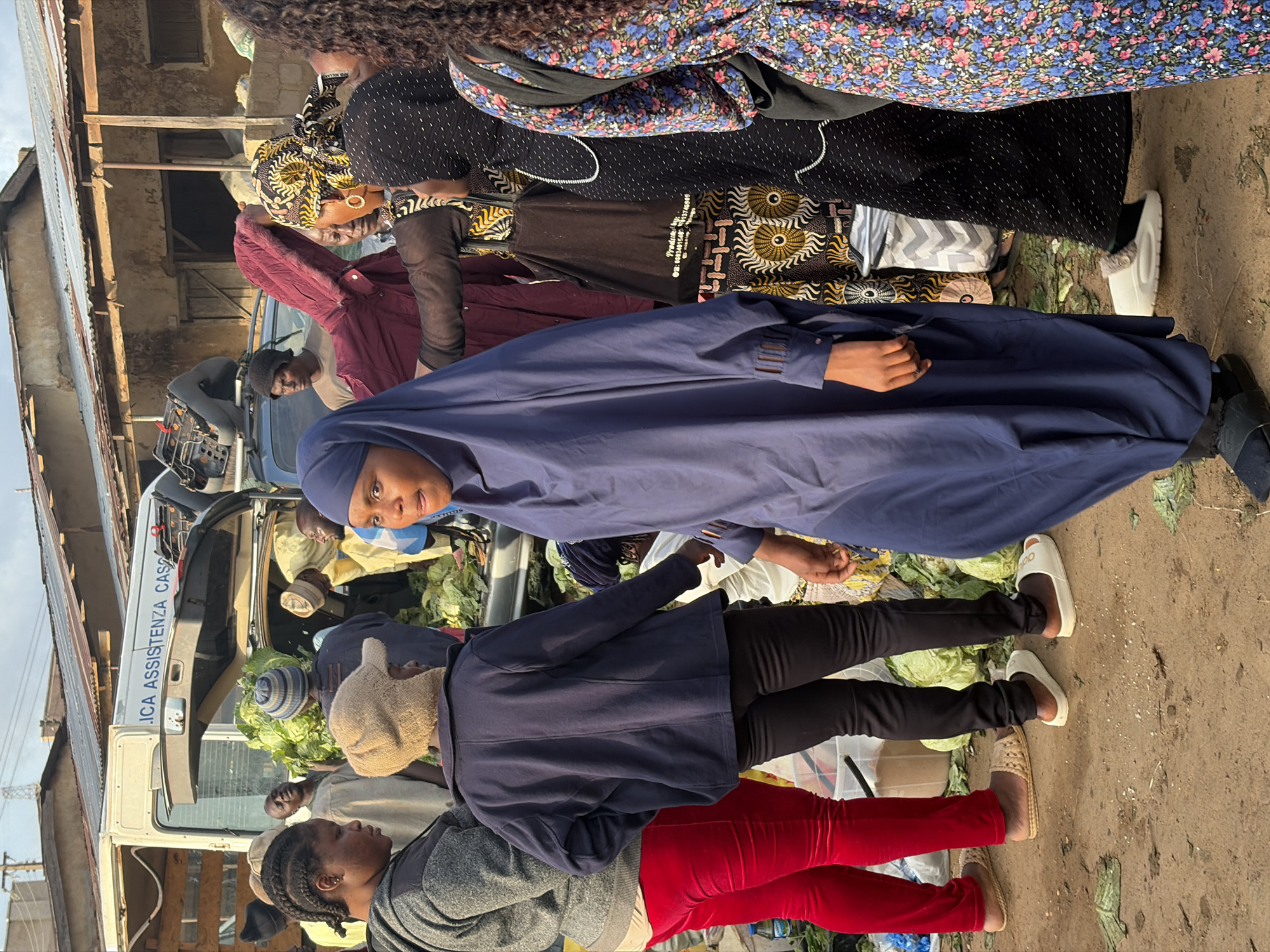 A group of people are gathered around a truck filled with produce, possibly at a market. The truck, which has "ICA ASSISTENZA CAS" written on its side, is a focal point. Several individuals are dressed in traditional Nigerian attire, including headscarves and brightly patterned fabrics. One person in the foreground is wearing a dark blue hijab and a matching long coat, with dark pants and white slides. Another person to their right is in red velvet pants and a gray top, also wearing sandals.
The scene appears to be outdoors, with a dirt ground covered in some scattered debris. The lighting suggests it might be daytime, possibly late afternoon given the angle of shadows, though the sky is not clearly visible. The people are engaged in what seems to be a transaction or interaction related to the produce. Some are looking at the truck, others are conversing. The atmosphere seems to be that of a busy market day. The people are mostly looking away from the camera, with the exception of the person in the blue hijab, who is looking directly at the camera. There is a generator visible on the truck.