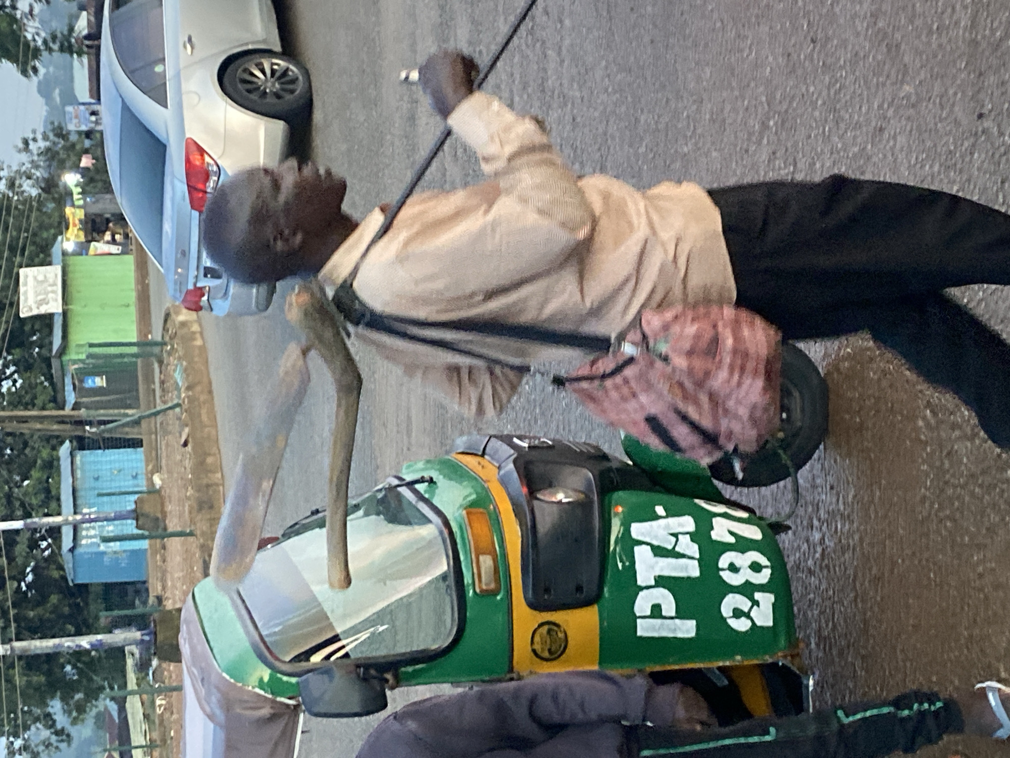 In Bukuru, Nigeria, a man walks beside a green and yellow auto-rickshaw. He carries a long, thick wooden pole across his shoulders, with a bundled bag slung over his right shoulder. The auto-rickshaw has "PTA" and "287" printed on its side in white lettering. In the background, a silver sedan is parked on the street, and various buildings and trees are visible. The scene appears to be taking place on a paved road with some puddles, suggesting recent rainfall. The lighting indicates it is daytime, possibly early morning or late afternoon, with soft, diffused light.