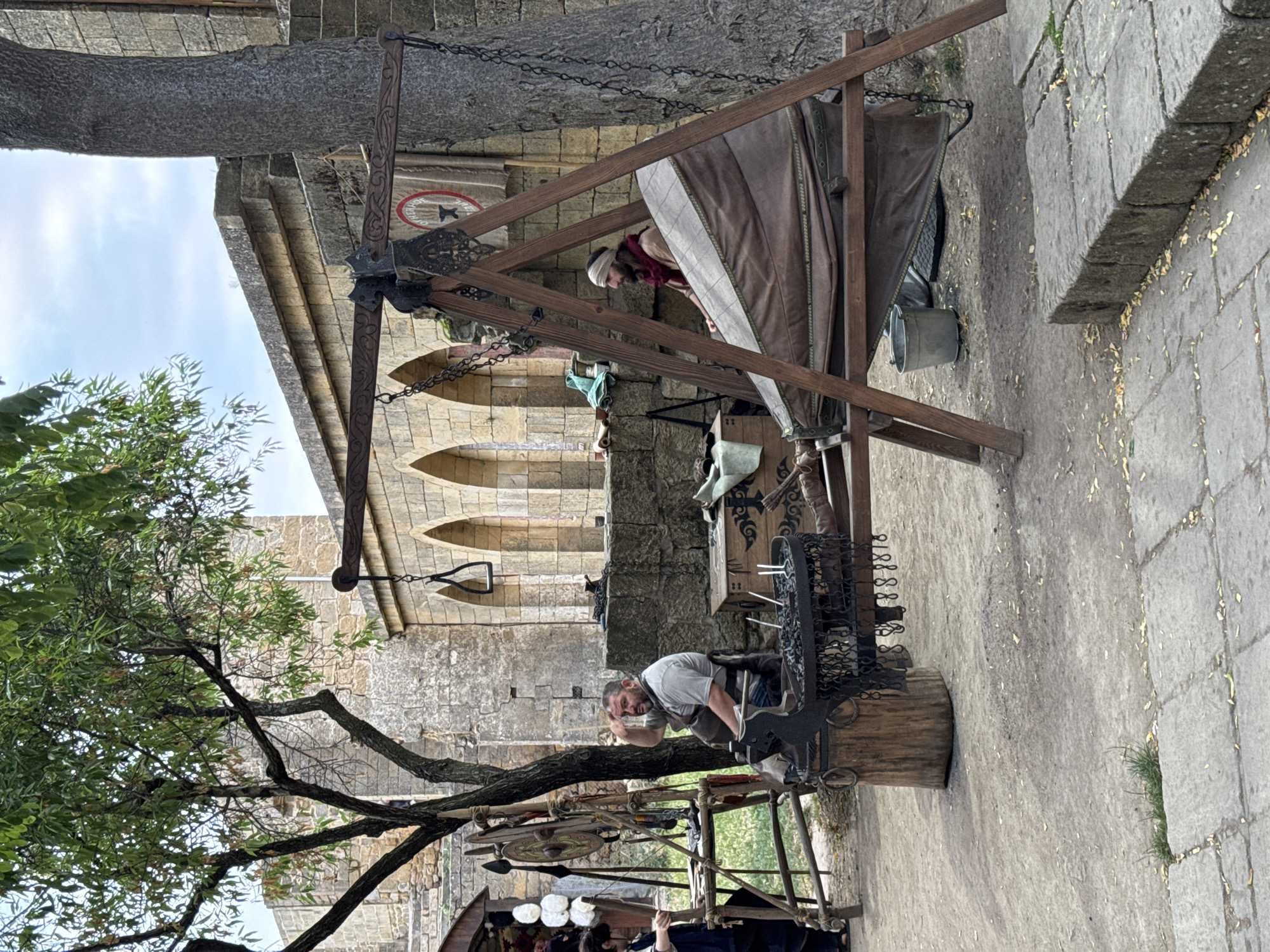 The image depicts a historical reenactment, likely a blacksmith's workshop, set against the backdrop of the ancient Naryn-Kala fortress in Derbent, Russia. In the foreground, a man with a beard, wearing a grey apron and shirt, sits on a wooden stool. He appears to be operating a bellows, which is part of a large, wooden structure with a chain mechanism. To his left, another man is visible, partially obscured, working at a table with various tools and materials, including spears and a shield. The environment consists of a dusty ground, stone walls, and arched openings characteristic of the fortress architecture. Several wooden chests and a metal bucket are also present. The scene suggests a demonstration of historical crafts and a glimpse into the past within a significant historical site.