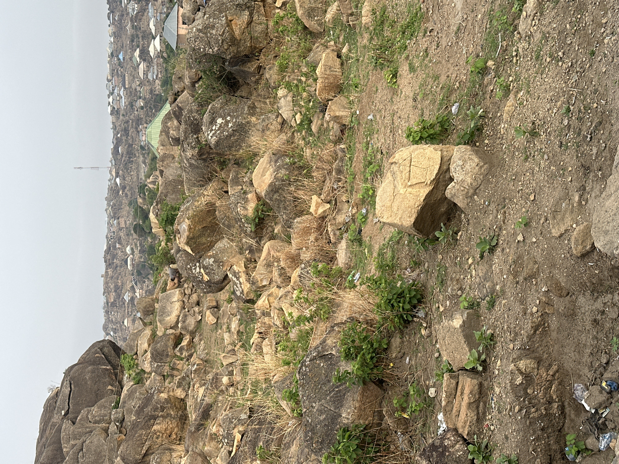The image presents a landscape in Jos, Nigeria, characterized by rocky terrain and sparse vegetation. In the foreground, large boulders and smaller rocks cover the ground, interspersed with dry grass and small green plants. The middle ground transitions into a more developed area, featuring rooftops of buildings clustered together. The background is hazy, obscuring distant details. The overall scene suggests a location on the outskirts of a city or town, where natural rock formations meet urban development.