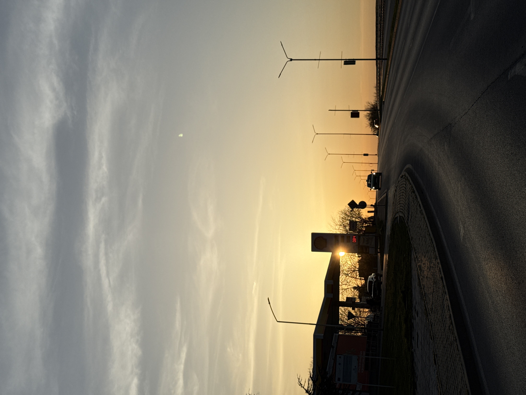 Captured at sunset in Dobrich, Bulgaria, a scene unfolds featuring a Shell gas station, silhouetted against the warm glow. The price display reads "0.84." A road curves beside the station, streetlights lining the path. Cars are parked at the gas station. A truck drives along the road, its silhouette distinct against the skyline. The sky transitions from light clouds to a golden hue, with the setting sun adding a halo. The scene exudes an atmosphere of dusk along a typical urban road.