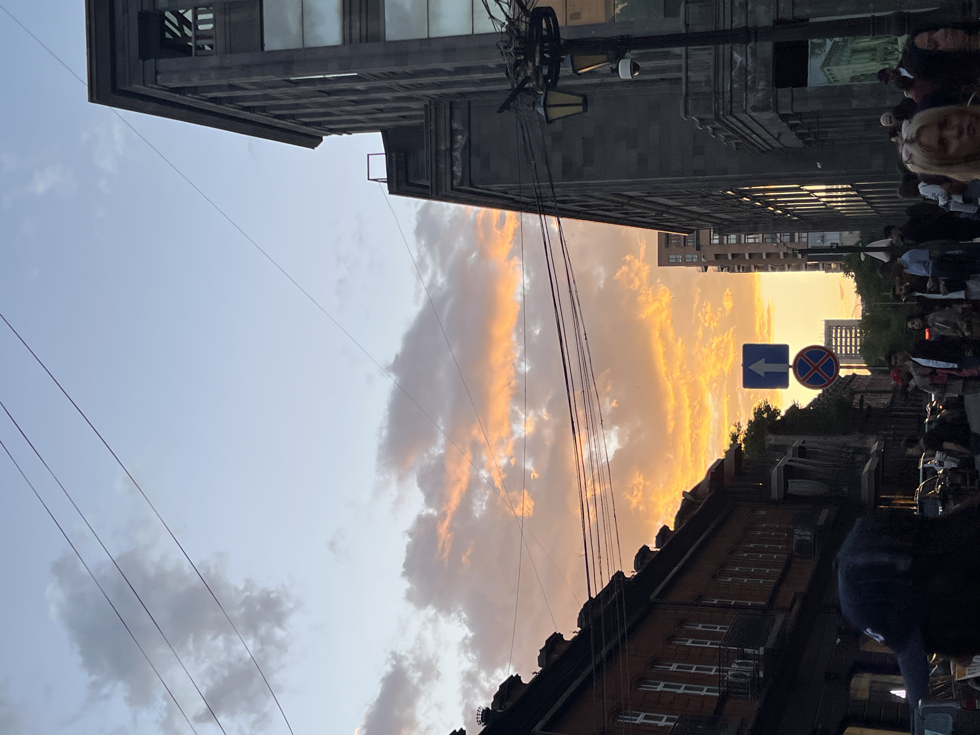 The image captures a Yerevan, Armenia street scene at sunset. Buildings line both sides of the street. Power lines stretch across the sky. The sky is filled with clouds, illuminated by the setting sun in orange and yellow hues. Pedestrians walk along the sidewalk. A blue and red traffic sign is visible.