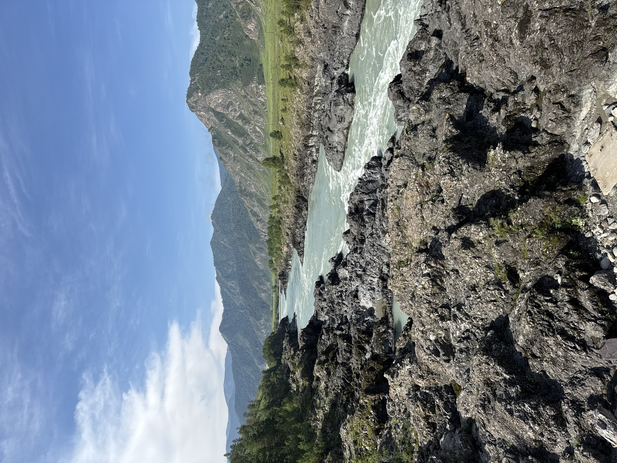 The image depicts a rugged, rocky landscape with a turquoise river flowing through it. The river is turbulent, with white water indicating rapids. On either side of the river are steep, rocky slopes covered in sparse vegetation, including trees. In the background, there are rolling mountains covered in green forests. The sky is blue with scattered white clouds. The scene appears to be natural and undeveloped. The location is identified as the Katun River in Edigan, Russia. There are no people or animals visible in the image, and no actions or interactions are taking place. The environment is characterized by its natural beauty and rugged terrain.