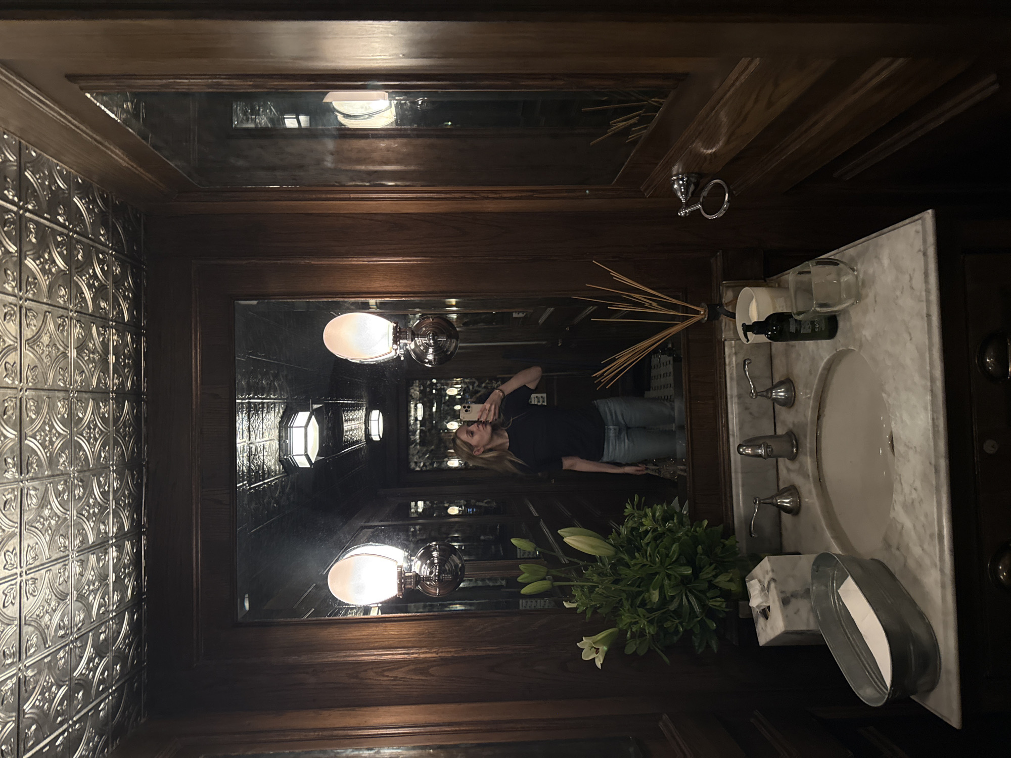 The image shows a dimly lit bathroom interior, likely taken in Mexico City based on the reflection in the mirror. A woman is seen in the mirror, holding a phone and taking a selfie. The bathroom has dark wood paneling, a decorative metal-tiled wall, and a marble sink. There are toiletries on the sink, along with a vase of flowers. Light fixtures are visible in the mirror reflection, suggesting a long, narrow space.