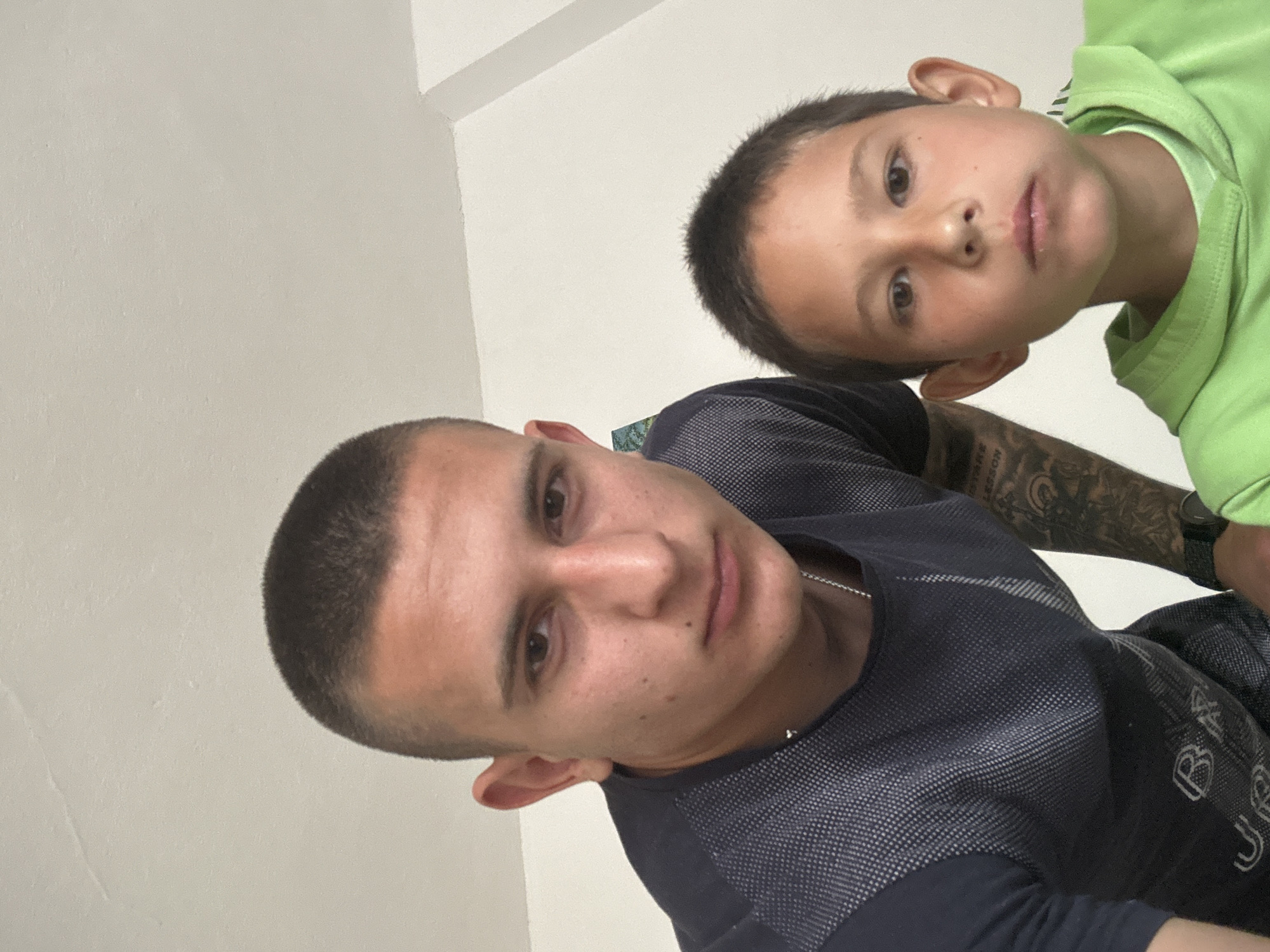 A man and a boy are posing for a close-up photograph, likely a selfie. The man has short, dark hair and is wearing a dark gray t-shirt with a textured pattern. Tattoos are visible on his left arm. The boy is positioned above the man and is wearing a bright green shirt. The background appears to be a plain white wall. The location is Kalchevo, Bulgaria, but there are no features in the image to confirm this.