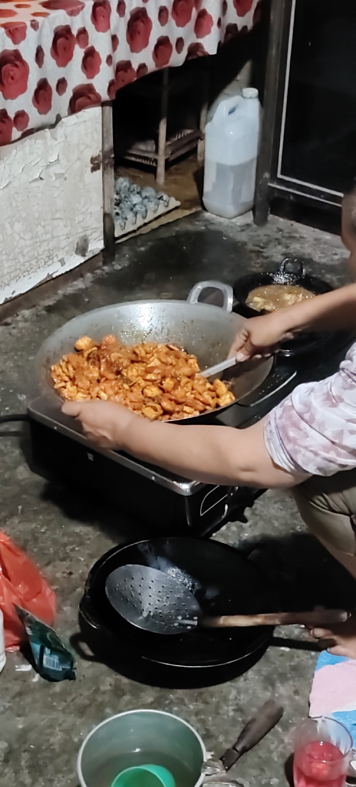 The image captures an indoor cooking scene in Dayah Tuha, Indonesia. A person, visible from the torso downwards, wearing a light-colored patterned shirt and dark trousers, is actively cooking. Their hands are positioned over a large, silver wok filled with numerous pieces of reddish-brown food, likely meat or poultry coated in sauce, resting on a black portable electric or induction cooktop. The individual uses a white spatula to stir the contents of the wok. To the right, a smaller black pan on another burner contains a lighter brown, saucy preparation.
The immediate foreground displays a red plastic bag, a green packaged item, a silver pot containing a green plastic cup, and a clear glass with a red beverage, possibly with ice or fruit, positioned on a concrete or tiled floor. A large black pan holding a metal slotted spoon with a wooden handle is also on the floor.
The background includes a wall or cabinet with peeling white paint and a small dark hinge. Above this, a table is covered with a white tablecloth featuring a recurring red rose pattern. Beneath the table, a wooden stool, a white tray containing multiple small, rounded, light-colored objects, and a tall, translucent white plastic jug filled with liquid are visible. A dark vertical beam or door frame stands to the right, beside a dark, reflective surface.