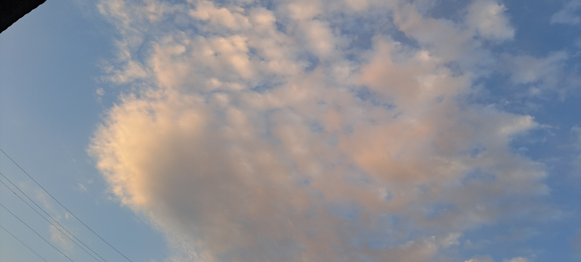 A wide-angle, eye-level shot depicts a pale blue sky filled with scattered, wispy clouds tinged with soft pink and orange hues, suggesting either sunrise or sunset. A dark, angular object, possibly the edge of a building or a roofline, occupies the upper left corner of the frame. Faint, parallel black lines, likely power lines or telephone wires, stretch diagonally across the lower left quadrant of the sky. The overall scene is an outdoor, atmospheric view without any visible people or specific urban landmarks within the city of Bash-Kaiyndy, Kyrgyzstan.