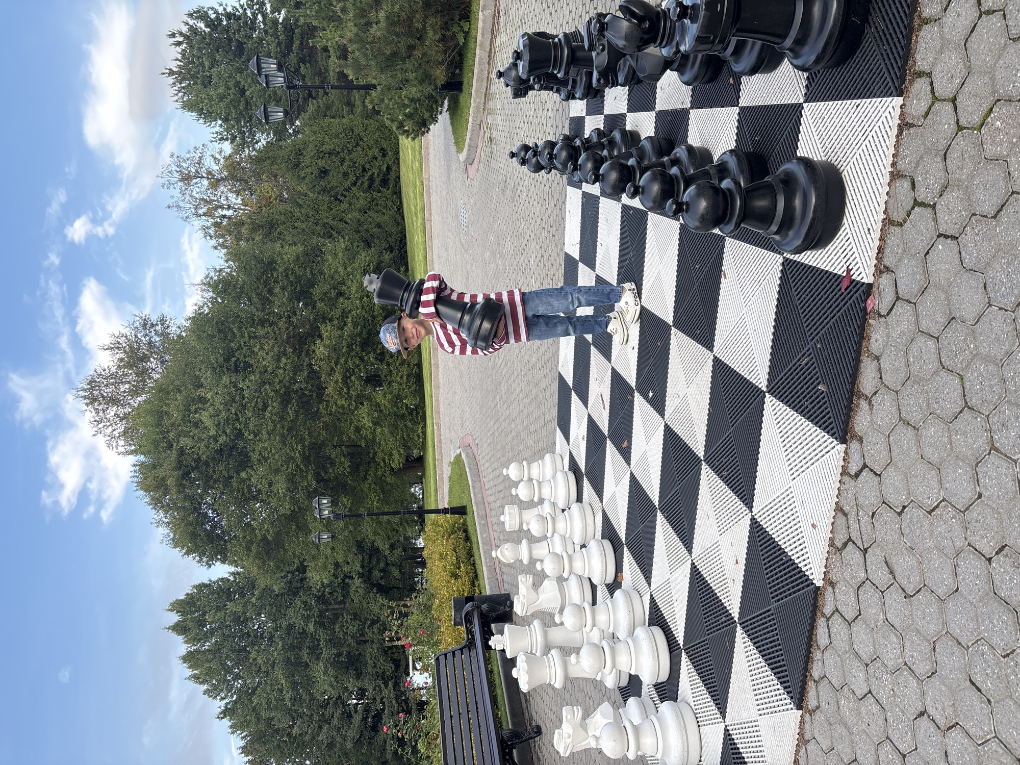 A young boy stands on a giant chessboard in Roždestveno, Russia, holding a black king chess piece. The boy is wearing a striped shirt, jeans, and a baseball cap. The chessboard is set up outdoors on a paved surface with hexagonal stones. The chess pieces are oversized, with white pieces on one side and black pieces on the other. In the background, there are trees, a park bench, and lampposts. The sky is blue with scattered clouds.