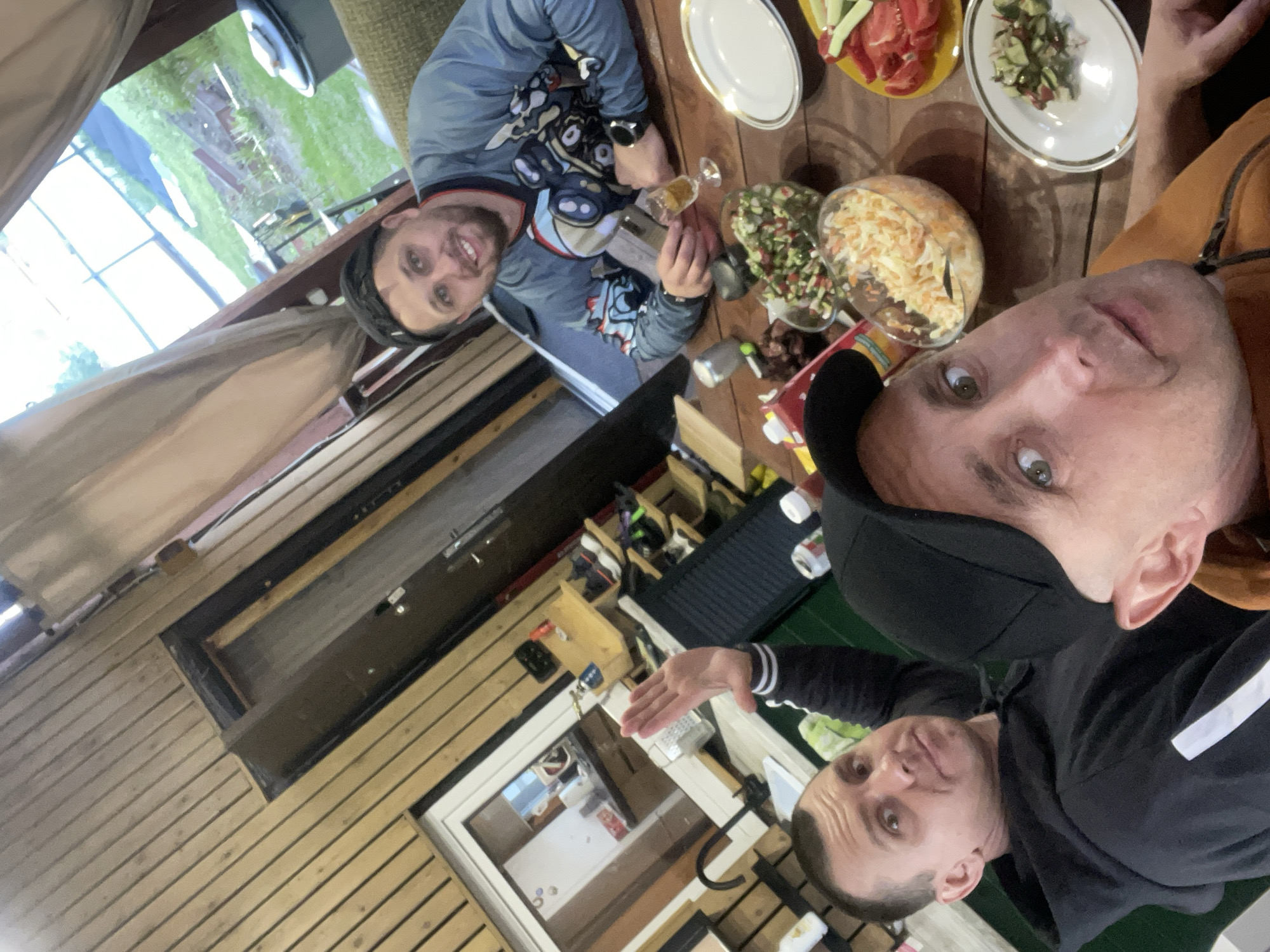 The image appears to be a selfie taken at a table laden with food. Three people are visible. One man wears a blue patterned shirt and a hat, holding a small glass. Another man, closest to the camera, wears a black baseball cap and an orange jacket. A third man is partially visible in a black hoodie. Plates of salad, sliced vegetables, and what appears to be coleslaw are on the table. The setting seems to be a covered patio or deck with wooden walls and a window. The location might be Bibikha, Russia.