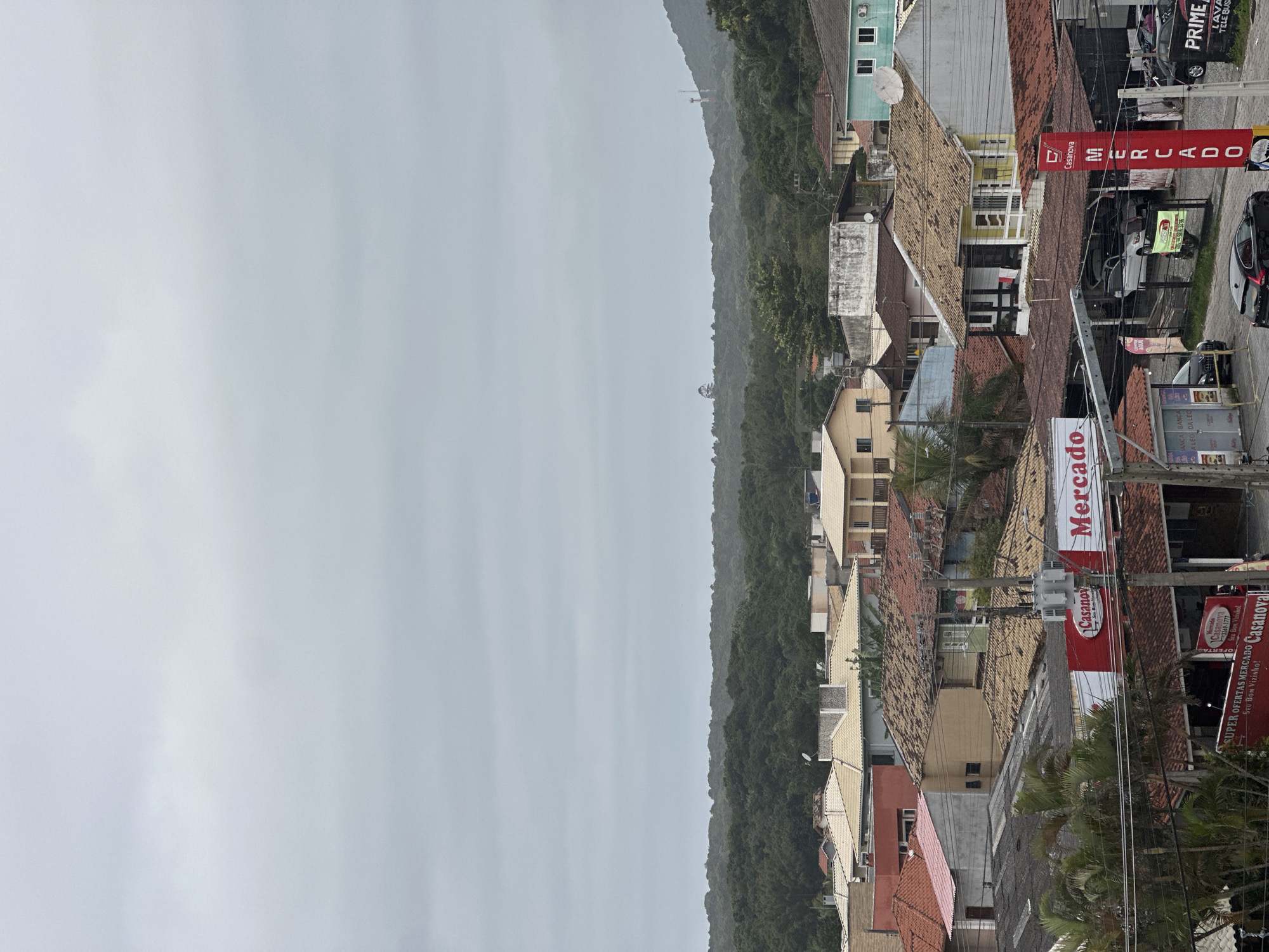 This is an aerial view of a densely packed urban area with a lush, green forest in the background. The foreground features a collection of buildings with tiled roofs of various colors and conditions, indicating a mix of residential and commercial properties. One prominent building displays a red and white sign that reads "Casnova" and "Mercado," suggesting a marketplace or supermarket. Another sign, also red and white, says "Mercado" vertically. There are several parked cars visible on the street and in what appears to be a parking lot. The sky is overcast and gray, creating a muted lighting condition for the scene. The architecture suggests a warm climate, possibly in Brazil, and the presence of a "Mercado" and distinct Brazilian signage confirms this is likely Florianópolis, Brazil. The dense foliage indicates proximity to a natural reserve or coastal forest common in Florianópolis.