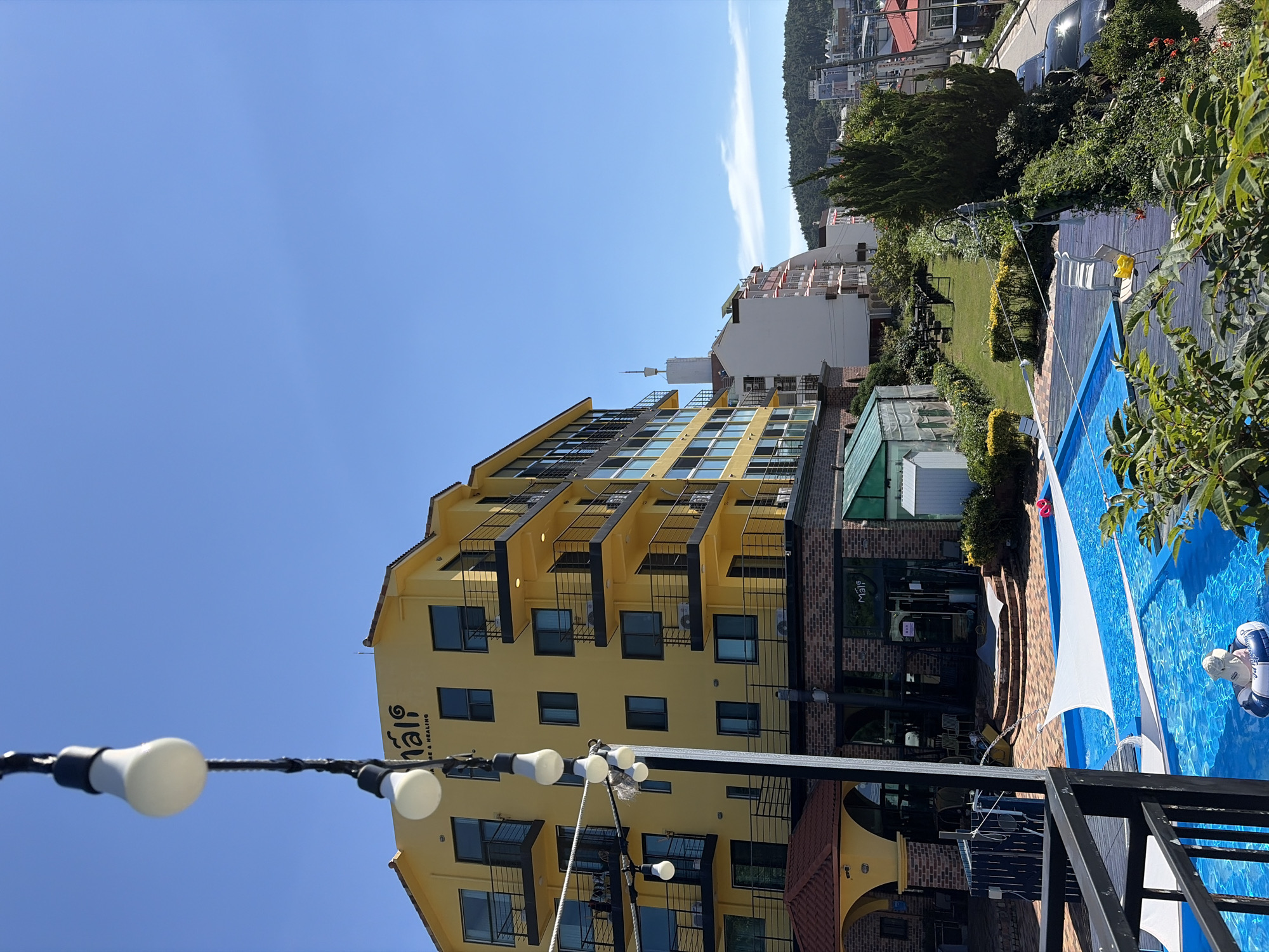 This image shows a view of a hotel building and a swimming pool, likely taken from a higher vantage point. The hotel is a multi-story building with a yellow facade and black balconies. A sign on the building reads "Mali Cafe & Healing". In front of the hotel, there is a bright blue swimming pool with white shade sails. A child in a white hat is floating on an inflatable ring in the pool. Adjacent to the pool is a grassy area with some trees and bushes. In the background, there are more buildings and a hillside covered with trees. The sky is clear and blue. The location is identified as Mohang-ri, South Korea.