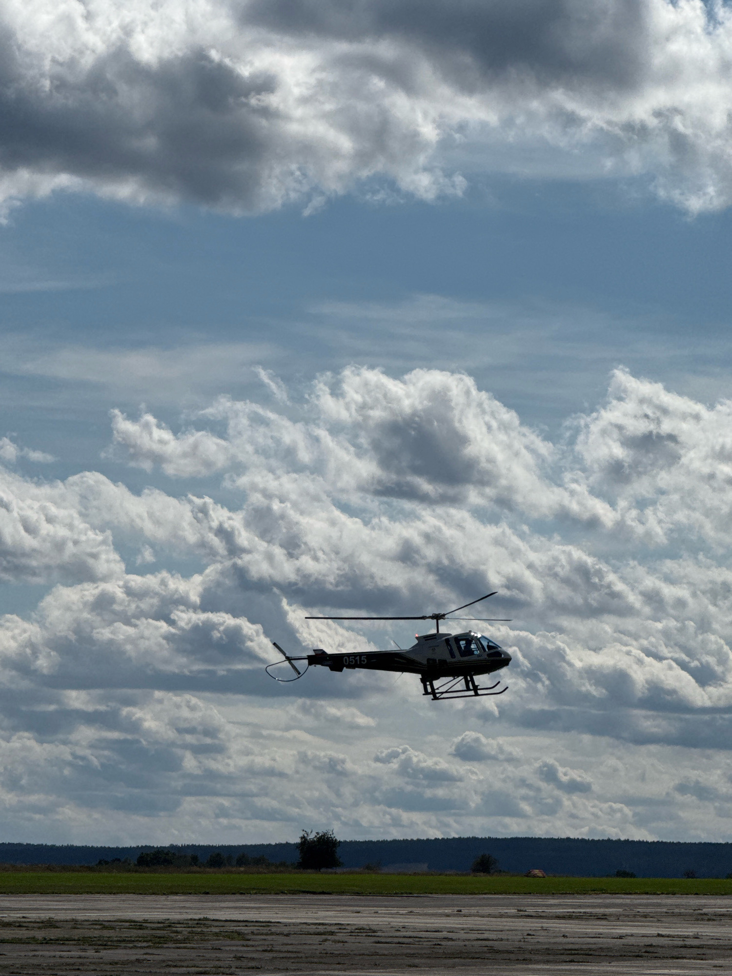 A helicopter, identified by the markings "0515" on its side, is in flight against a sky filled with dramatic, layered clouds. The helicopter is dark-colored with a lighter fuselage and tinted windows. It appears to be a light utility or surveillance helicopter. Below the helicopter, a flat landscape is visible, featuring a green field bordered by a dark, tree-lined horizon. In the foreground, a paved or gravel surface, possibly a runway or an open area, is present. The overall scene suggests an outdoor setting, likely an airfield or open land near Cheb, Czechia. The lighting indicates it is daytime, with sunlight filtering through the clouds. There are no people visible in the frame.