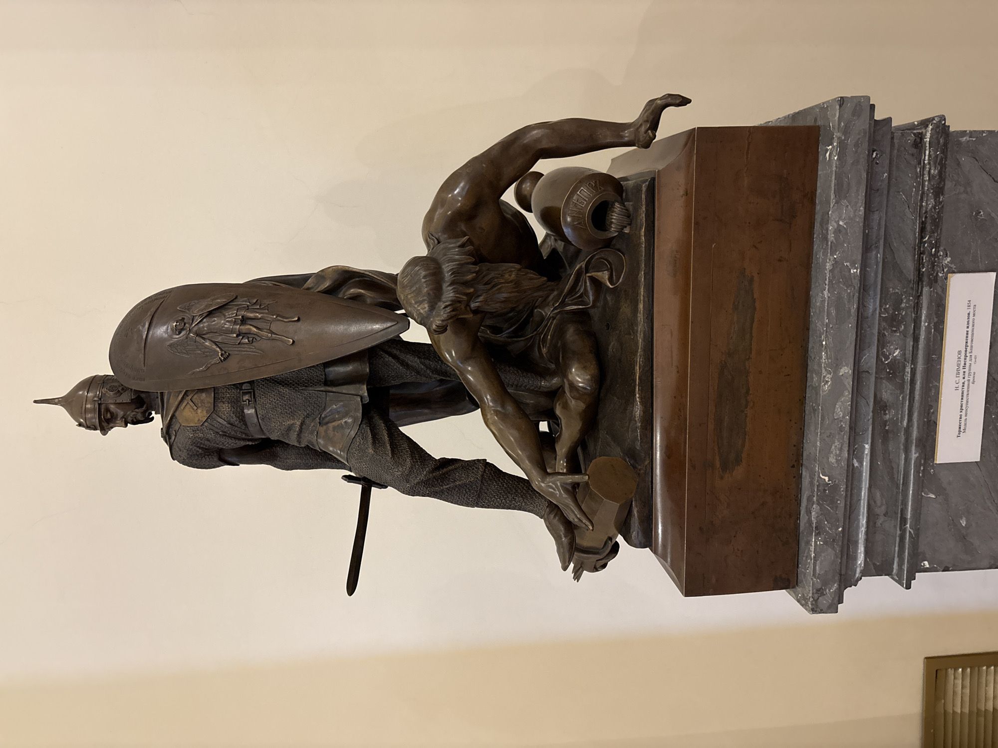 The image depicts a bronze statue by N.S. Pimenov, titled "The Triumph of Christianity, or the Overthrow of Idols, 1854." The statue shows a knight wearing a helmet and chainmail, holding a shield with a depiction of an angel on it, battling a shirtless figure who is struggling to hold a pot. The statue is mounted on a marble and wood base, and a sign identifies it as a model for a group that was not ultimately implemented for the Blagoveshchensky Bridge in Saint Petersburg, Russia.