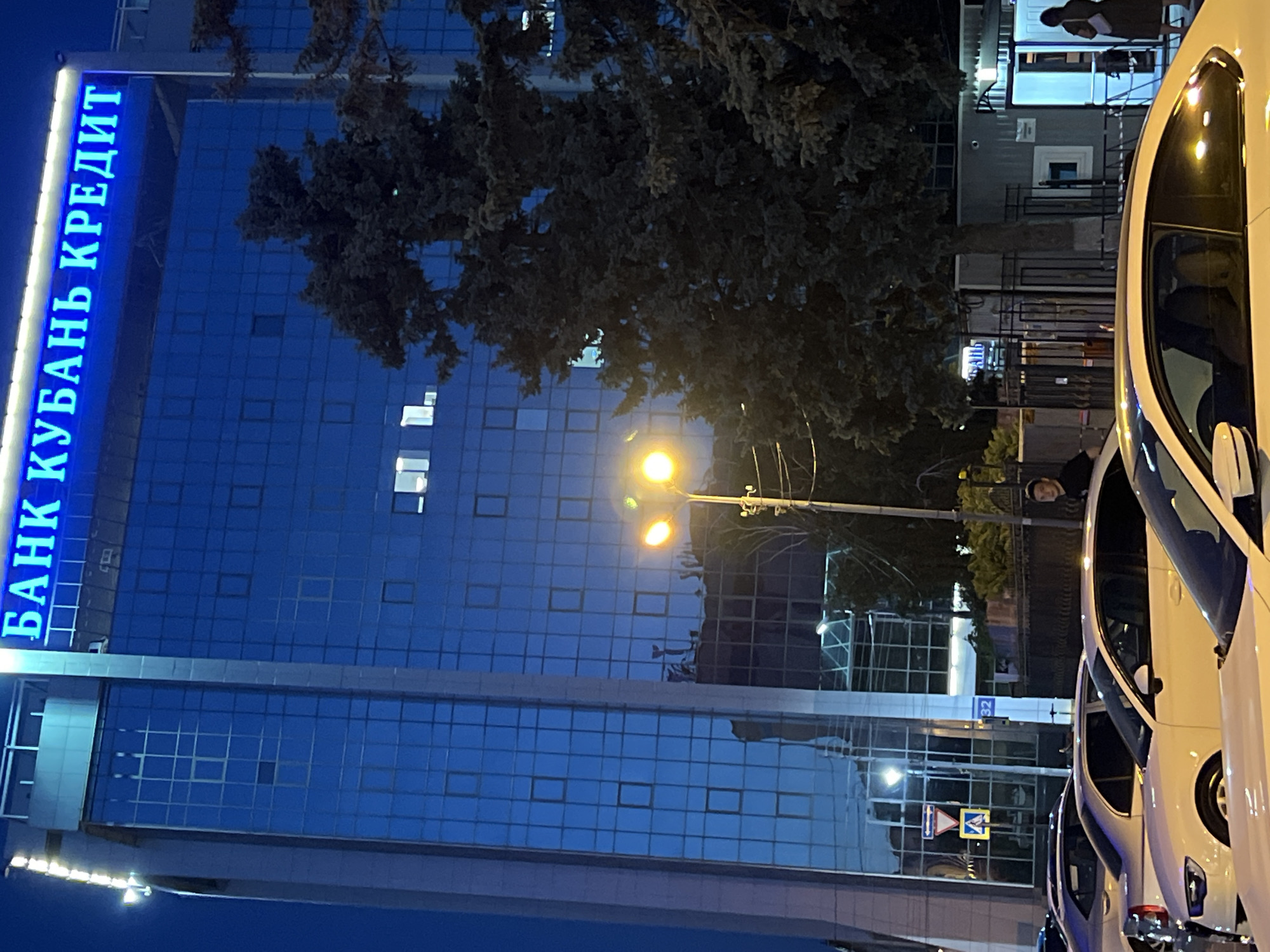 This is a vertical, outdoor, night-time photograph of a modern building with a blue glass facade. The building is illuminated, and neon blue lettering on the left side spells out "БАНК КУБАНЬ КРЕДИТ" (BANK KUBAN CREDIT). Several windows on the building are lit, suggesting interior activity. A streetlamp with a bright yellow light illuminates the scene.
In the foreground, the side of a white car is visible, as is the reflection of a person in its window. Behind the car, there appears to be another person standing near the entrance of a building. Further back, more vehicles and parts of buildings are visible. A tree with dark foliage partially obscures the view of the building.
The overall atmosphere is urban and nocturnal. The illuminated signage and building suggest a commercial or financial establishment. The presence of cars indicates a public area, possibly a street or parking lot. The clear sky and artificial lighting suggest no adverse weather conditions. The image is shot from a low angle, looking upwards towards the building.