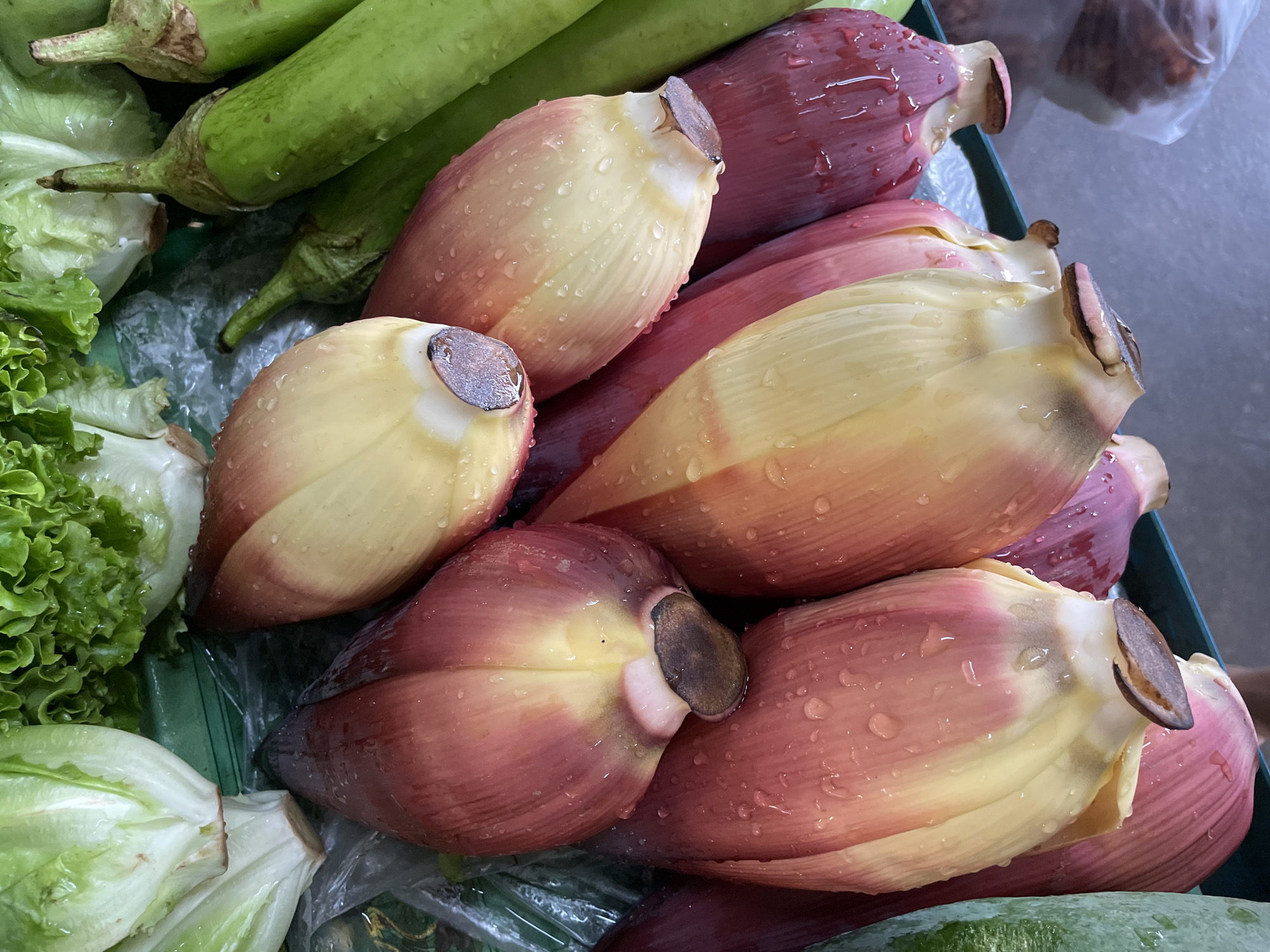 An overhead, close-up shot shows a collection of banana blossoms, some with purple outer layers and pale yellow inner layers, others with predominantly yellow inner layers and hints of pink, all glistening with water droplets. The blossoms are arranged in a pile, with their stems facing the viewer. In the background, to the left, are heads of lettuce with crisp green leaves. To the right, a row of long, green eggplants is visible. The scene appears to be a market stall in Nong Prue, Thailand, with the items likely for sale. The surface beneath the produce appears to be a green plastic tray.