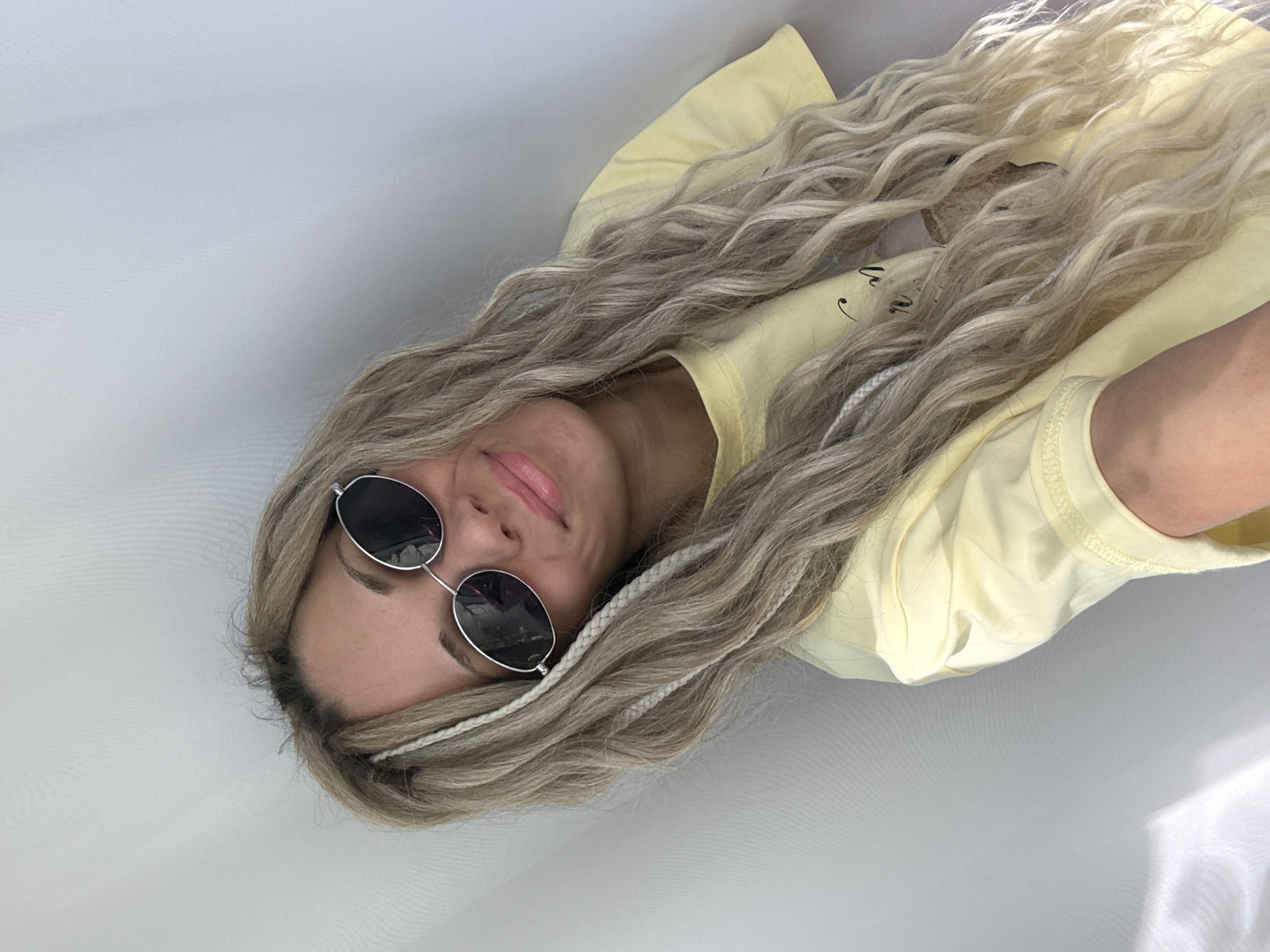 A woman with light-blonde, wavy hair, some braided, lies supine on a white surface. She is wearing round, black sunglasses with silver frames and a pale-yellow t-shirt. The image is taken from above, slightly angled. The background is predominantly white. No specific details indicate the location as Rusanovo, Russia.