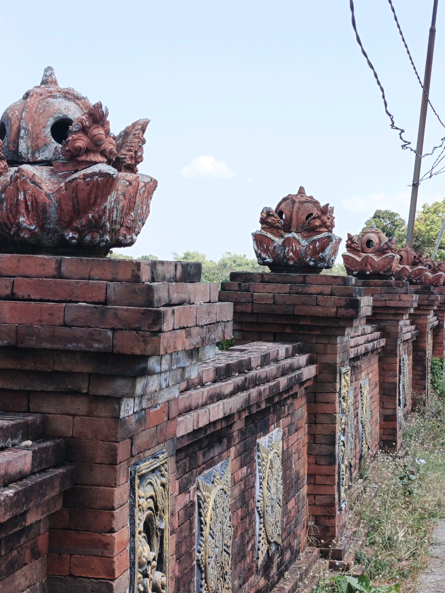 The image depicts a decorative brick wall with ornate sculptures atop it, situated in a rural or suburban area, possibly in or near the village of Panggih, Indonesia. The wall is constructed from reddish-brown bricks and features relief carvings on some of its vertical pillars. The sculptures, also made of brick or a similar material and painted a faded red, resemble mythological creatures or stylized heads, each with distinct facial features and decorative elements. They are positioned at intervals along the top of the wall. The background reveals a lush green landscape with trees and vegetation under a pale blue sky with a few wispy clouds. Electrical wires are visible in the upper right portion of the frame, suggesting the presence of infrastructure. The overall scene conveys a sense of weathered artistry and a connection to local cultural aesthetics.