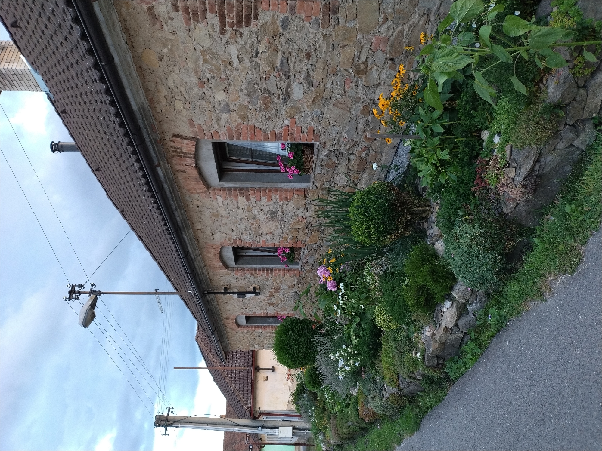 The image shows a street view of a stone-built house in the village of Dobev, Czechia. The house has a brown tiled roof and brick accents around the windows and corners. In front of the house, a well-maintained garden with various plants and flowers is visible, bordered by a low stone wall. A paved road runs along the side of the garden. Several utility poles with power lines and a street lamp are present in the background, indicating a rural or suburban setting. The sky is overcast with clouds. There are no people or animals in the image.