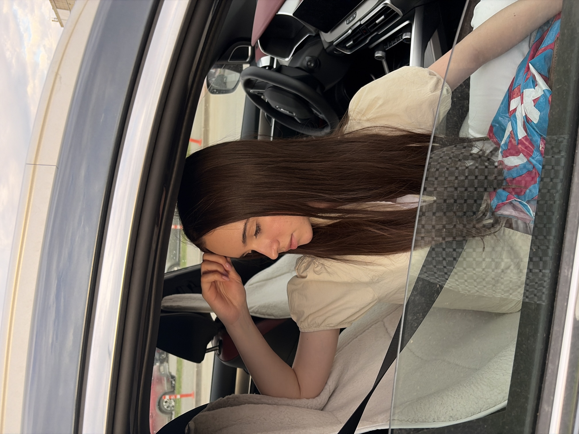 A young woman with long brown hair is sitting in the driver's seat of a car. She is wearing a light-colored top with puffy sleeves. Her head is resting on her hand, and she appears to be looking down. The car's interior includes a black steering wheel and dashboard. The exterior view through the car window shows a glimpse of what appears to be a road and possibly other cars, suggesting a city street or parking lot environment. Due to limited visual information, it is impossible to ascertain the location is in the city of Izhevsk, Russia.