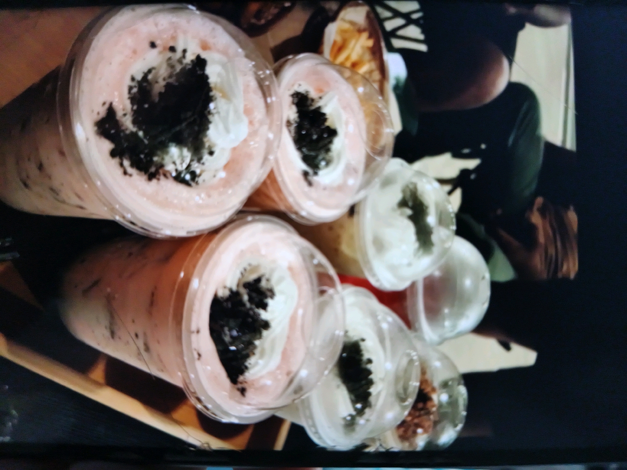 A collection of transparent plastic cups containing pink beverages and whipped cream, topped with crushed dark cookies, are arranged on a wooden surface. The beverages are blended, with visible ice particles. Some cups also contain a lighter colored liquid and what appears to be sprinkles. In the background, a person wearing a dark green shirt is partially visible, along with a portion of a container holding french fries. The scene is photographed from an overhead perspective, with the lighting creating reflections on the cups. The overall impression is that of a casual dining or takeaway setting. The location is not specified.