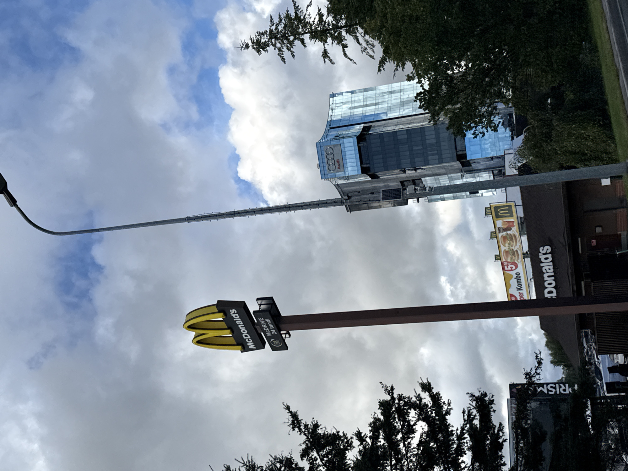 The image shows a cloudy day scene in Tallinn, Estonia, with a prominent McDonald's restaurant and its drive-thru sign in the foreground. A modern glass-fronted building, identified by an Audi logo, stands in the background. A tall lamppost is visible to the right of the McDonald's sign. The McDonald's sign is on a pole and displays the company's golden arches logo, along with text indicating "McDonald's" and "McDRIVE 24 tundi". A billboard advertising a "per Kombo" meal from McDonald's is also visible above the restaurant's roof. The restaurant itself appears to be a standard McDonald's structure with a dark roof and brown facade. The surrounding environment includes trees and a road in the lower right corner. Several cars are parked in what appears to be a parking lot behind the McDonald's. The sky is filled with a mix of dark and light grey clouds, with some patches of blue sky visible.