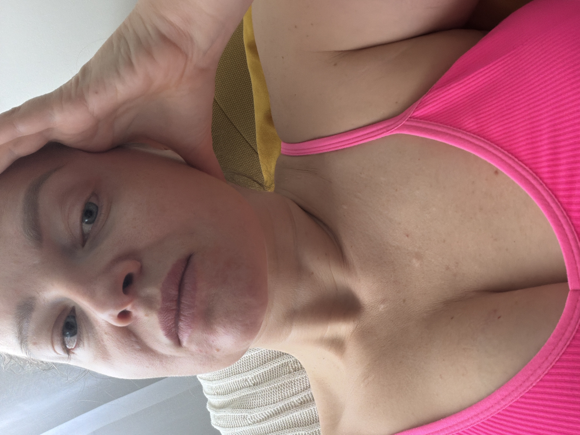 A close-up, low-angle selfie shows a light-skinned woman with gray eyes and light brown hair. She is wearing a ribbed, pink top with a rounded neckline. Part of her left hand is on her forehead. The background includes a mustard-yellow object and a beige textured surface. No information in the image suggests a location in Moscow, Russia.