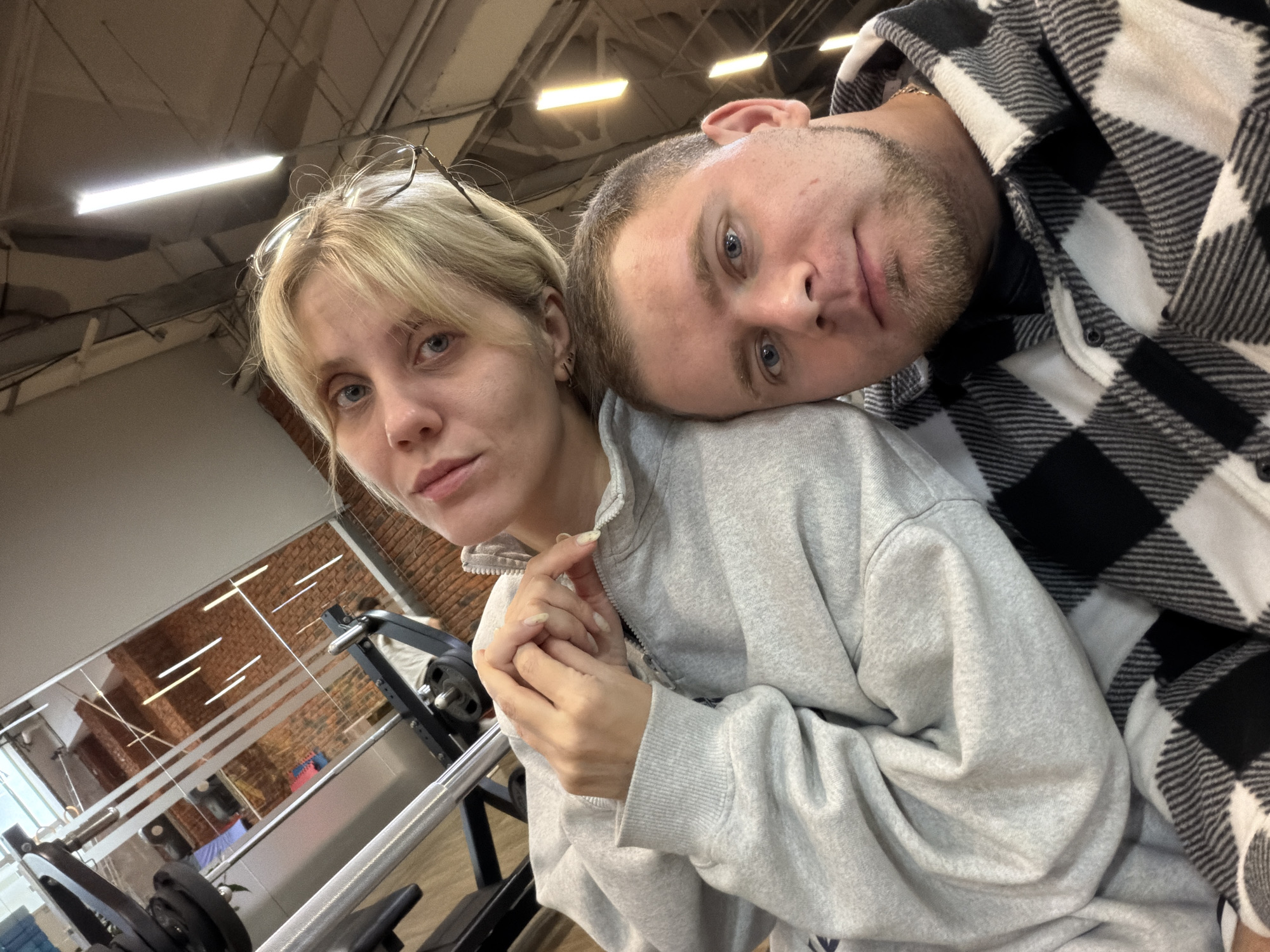 A selfie captures two individuals, a man and a woman, in what appears to be an indoor gym or fitness center in Chaikovsky, Russia. The image is taken from a slightly upward angle, focusing on the couple.
The woman, with blonde hair and bangs, is in the foreground, looking directly at the camera with a neutral expression and blue eyes. She wears a light grey zippered hoodie, and a pair of glasses rests on top of her head. Her hands are clasped near the zipper of her hoodie, revealing long, lightly polished fingernails.
The man is positioned behind and to the side of the woman, with his head gently resting on her shoulder or upper chest, conveying a sense of comfort or intimacy. He has short hair, light stubble on his face, and blue eyes, also looking directly at the camera with a neutral gaze. He is dressed in a black and white plaid hooded jacket or shirt, layered over a darker garment.
The background clearly identifies the setting as a gym. Exposed ceiling structures with visible pipes and rectangular fluorescent tube lights are overhead. Through a clear glass partition, a brick wall is discernible in the mid-ground, possibly indicating an adjacent area or a building exterior. Various pieces of gym equipment, including parts of a barbell and other exercise machines, are visible in the foreground and behind the couple, reinforcing the fitness environment. The overall lighting is artificial and bright. There is no visible text in the image.