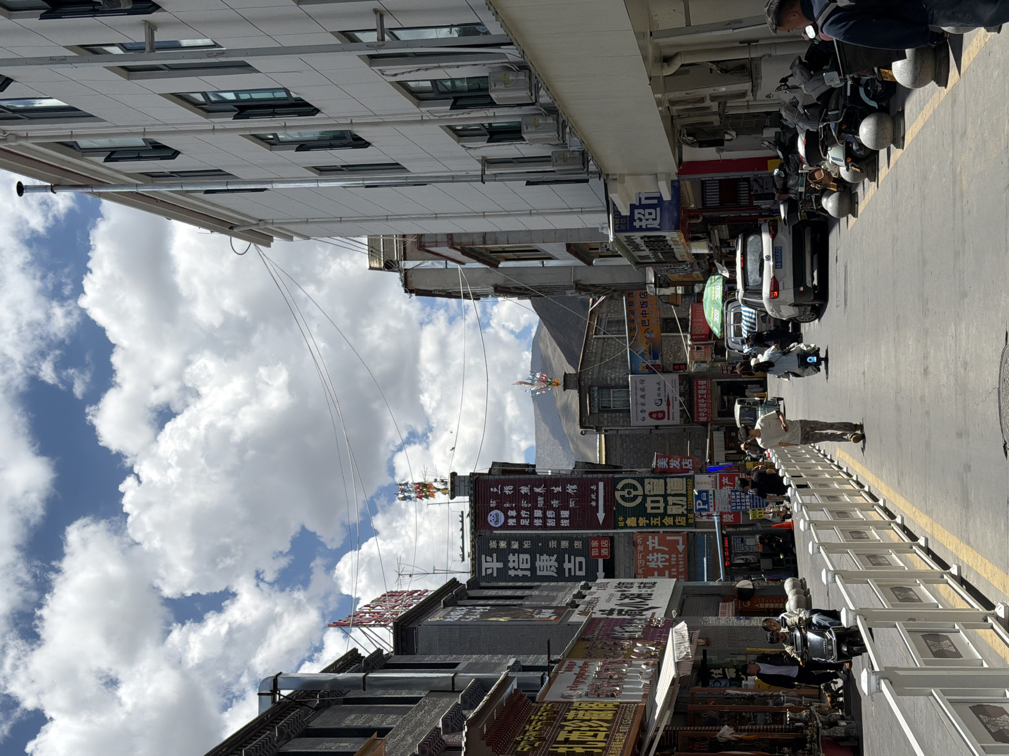 This image captures a lively street scene in what appears to be a bustling town or city, likely in Chengguan District, China, with mountainous terrain visible in the background. The photo is taken during bright daylight, indicated by the clear blue sky adorned with fluffy white cumulus clouds.
The street is lined with multi-story buildings of varying architectural styles, some appearing more modern with grid-patterned facades and numerous windows, while others have a more traditional look. Many of these buildings host a multitude of businesses, indicated by the array of colorful commercial signs hanging outside. A paved road, marked with a yellow dividing line, runs through the center, with a white, low barrier fence separating it from the sidewalk or storefronts on the right.
Several people are visible in the scene: a person walking on the street in the middle distance, another individual on the far right near parked vehicles, and people seated on or near motorcycles and scooters. A significant number of motorcycles and scooters are parked along the street, and a white car with the license plate "A-66420" is also visible further down. Overhead, a tangle of power lines and cables crisscrosses the street, adding to the urban feel. Traditional decorative elements, possibly prayer flags, can be seen atop some distant buildings, hinting at the cultural context of the region.
Visible text from the numerous signs includes "Three-Finger Health Preservation Hall," "Pintso Kangji," "Jiale Pai. Yun Hotel Home Store" (possibly a hotel), "Baixiang Family Hostel," "Bazhong Hotel," "Hair Salon," "Zhongxinyu Hardware Store," and a "Handmade Steamed Bun Shop." Other signs advertise "specialty products" such as "yak jerky," and Tibetan script (ཁྲོན་ཁྱད་རྫས།) is also visible, reinforcing the location's cultural identity.