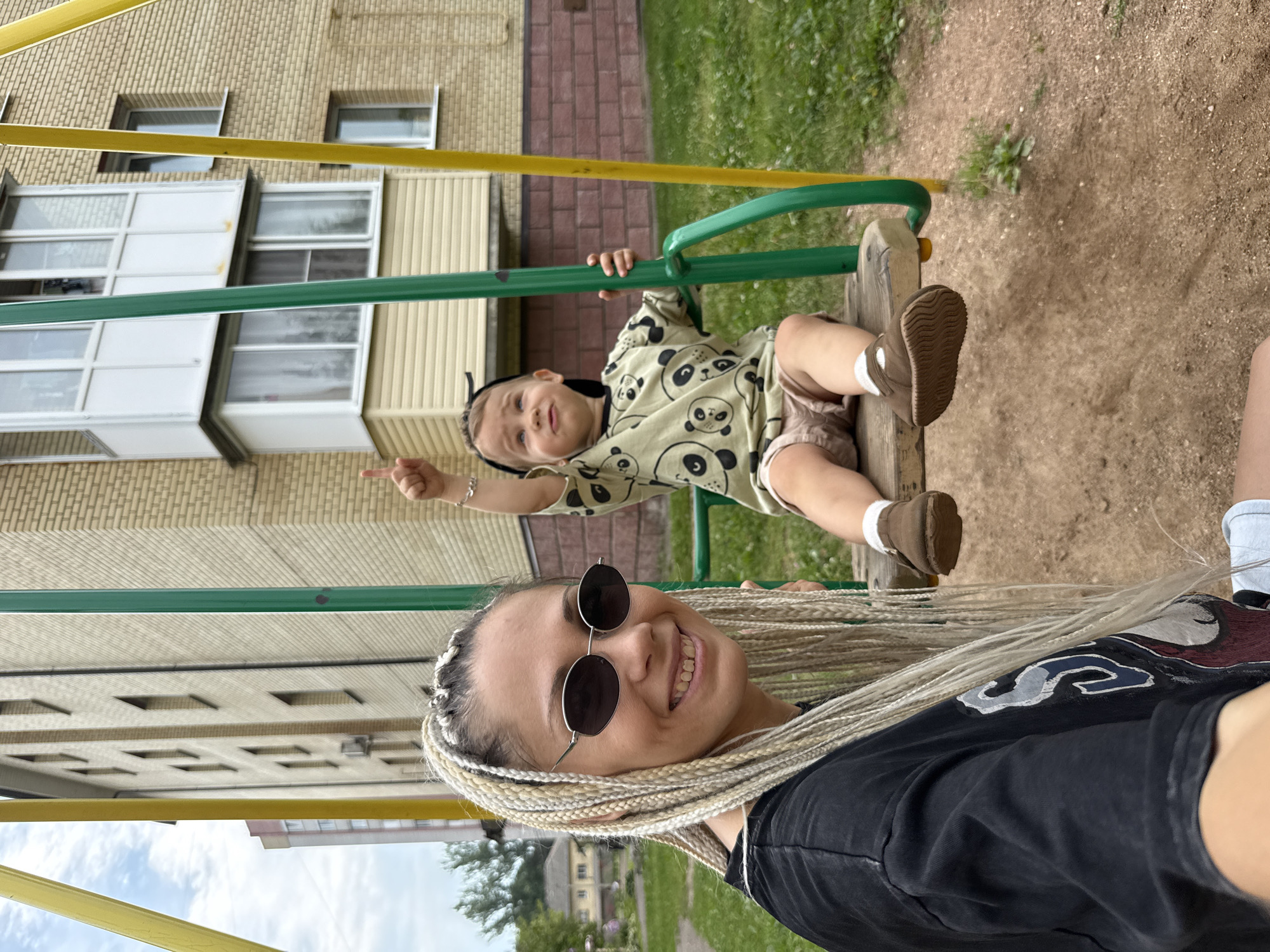 In Rusanovo, Russia, a woman with blonde braided hair and sunglasses smiles at the camera, while a young child on a swing behind her points upwards with their right hand. The child wears a light green shirt with a panda print and light brown shorts. They are sitting on a wooden swing seat suspended by green and yellow metal poles, situated on a playground with a sandy ground and grassy areas. A multi-story brick building is visible in the background, with windows and balconies. The scene is outdoors, under a cloudy sky.